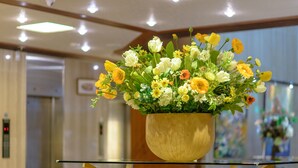 Um vaso de madeira com um arranjo floral sobre uma mesa de vidro num saguão de hotel.