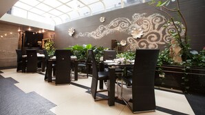 Uma sala de jantar moderna com teto alto e luz natural, com cadeiras e mesas pretas, arte decorativa na parede e plantas dentro de casa.
