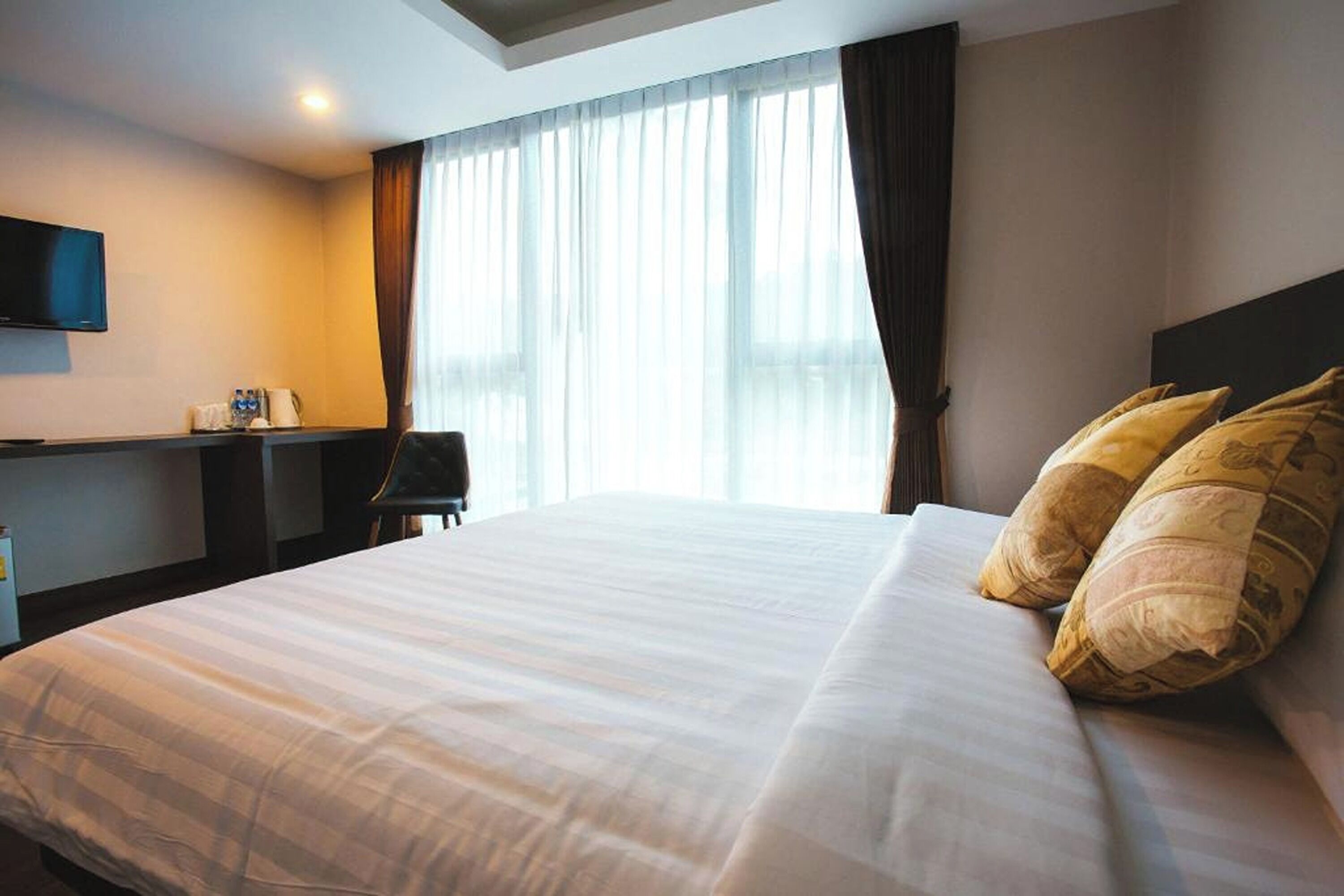 A hotel room with a large bed, a flat-screen TV mounted on the wall, a desk with a chair, and a window with curtains.