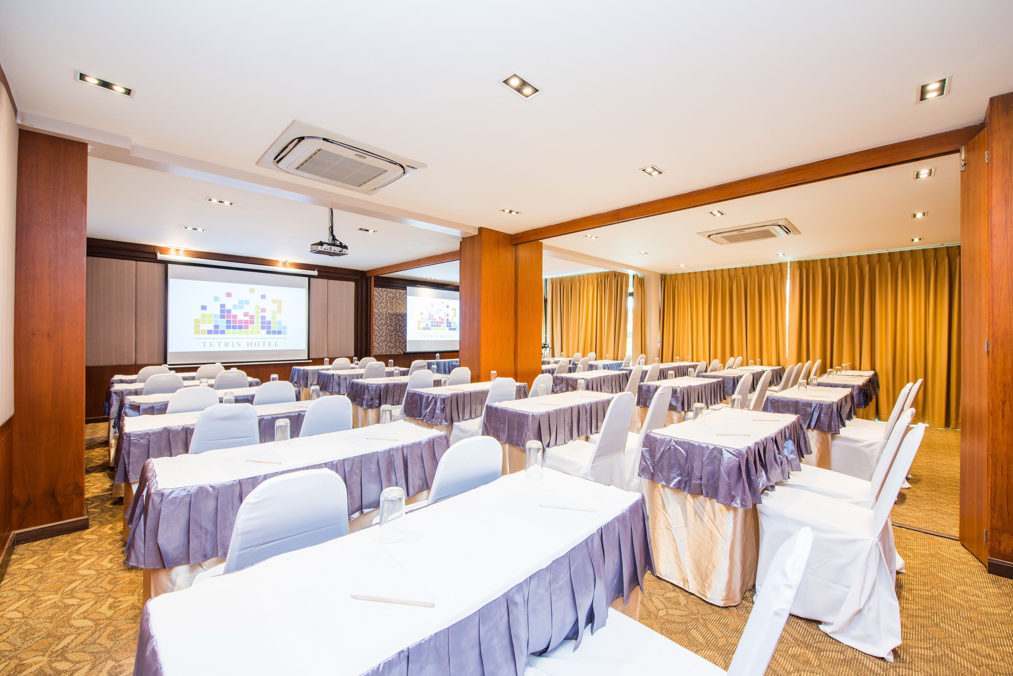 Un salón de conferencias con filas de mesas y sillas, una pantalla de proyector y una pantalla para presentaciones.