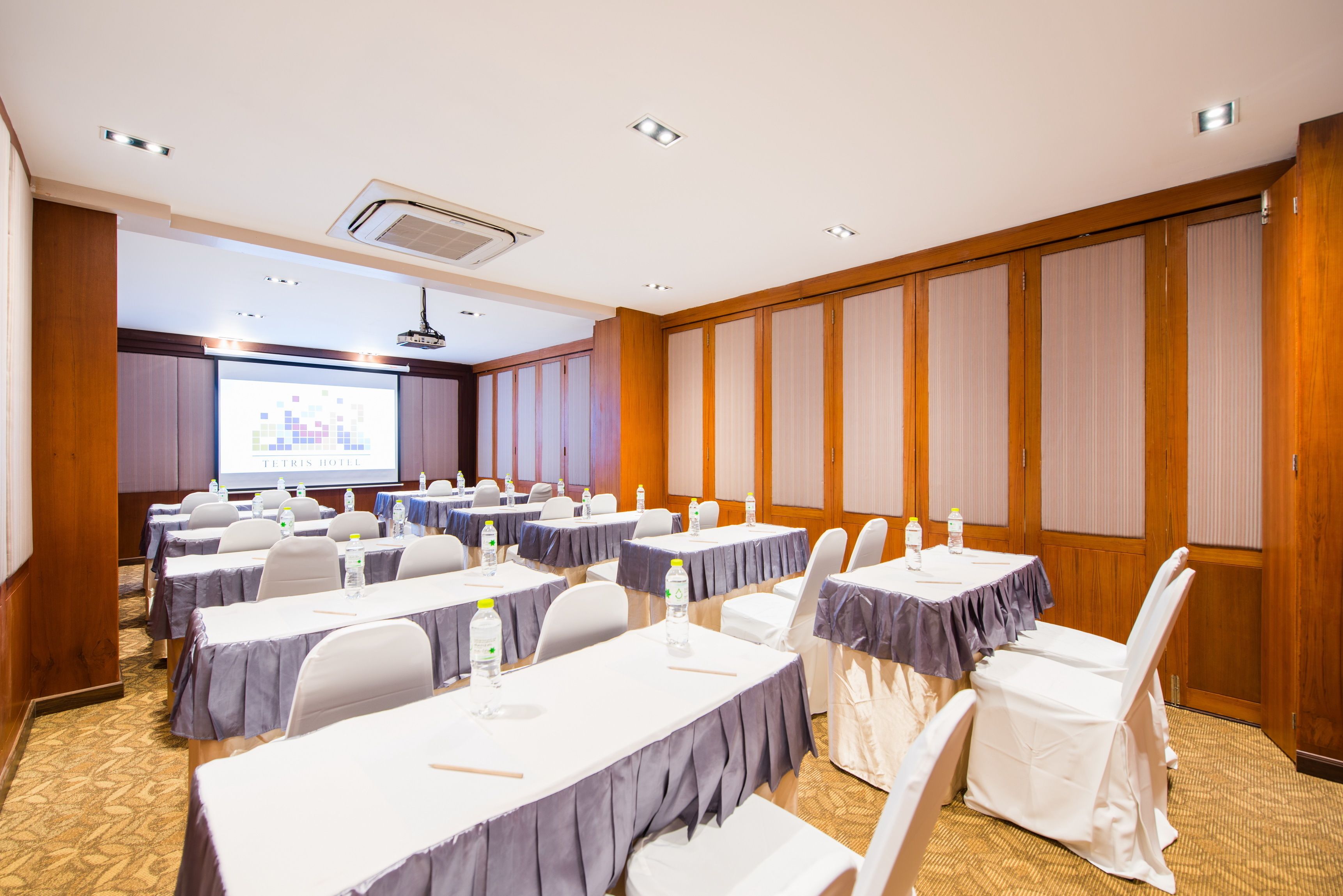Un salón de conferencias con varias mesas, sillas y una pantalla de proyector mostrando 'ELTRIS HOTEL'.