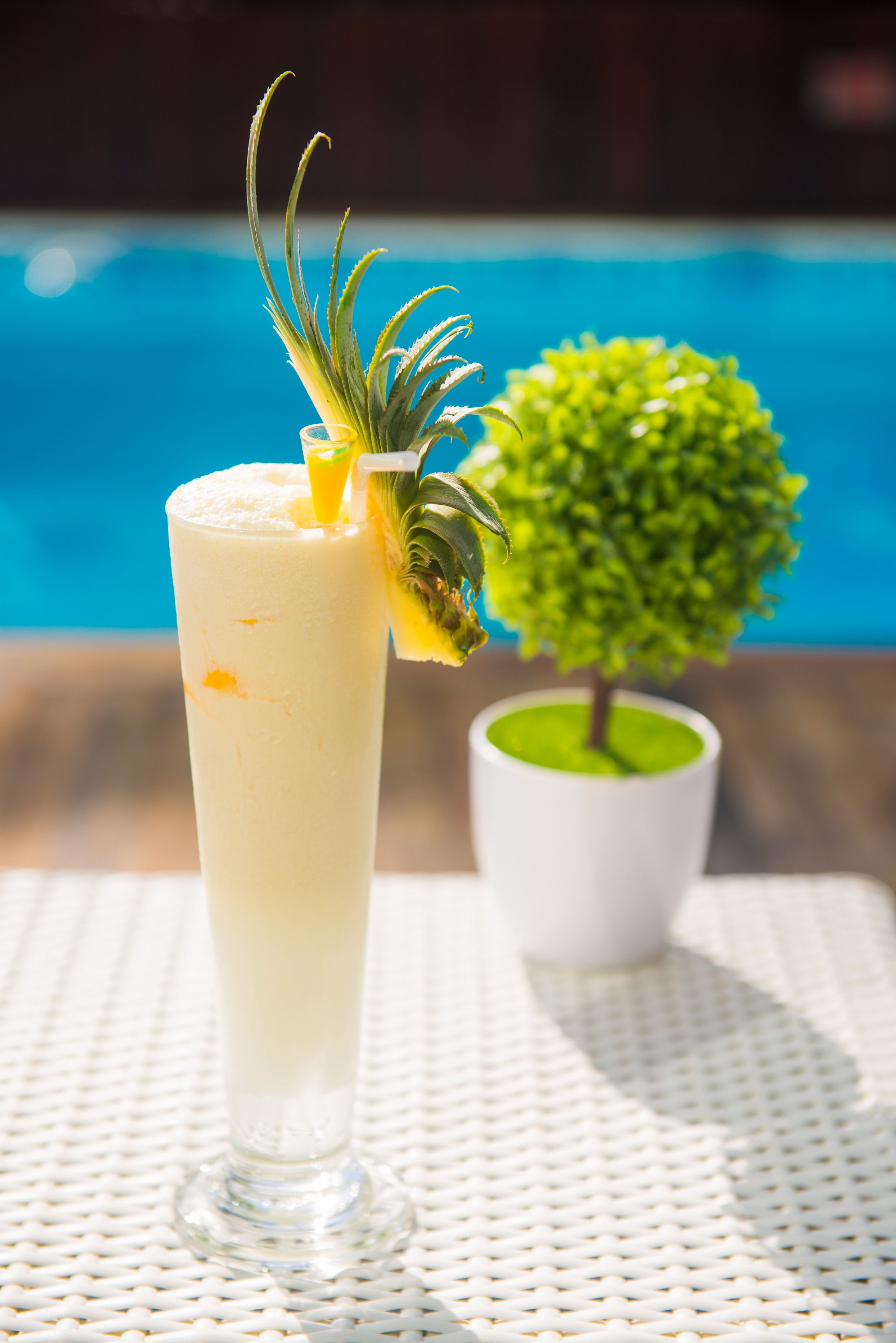 A tall glass with a creamy beverage, garnished with a pineapple and a decorative leaf, placed on a white textured surface. A small potted plant is visible in the background.