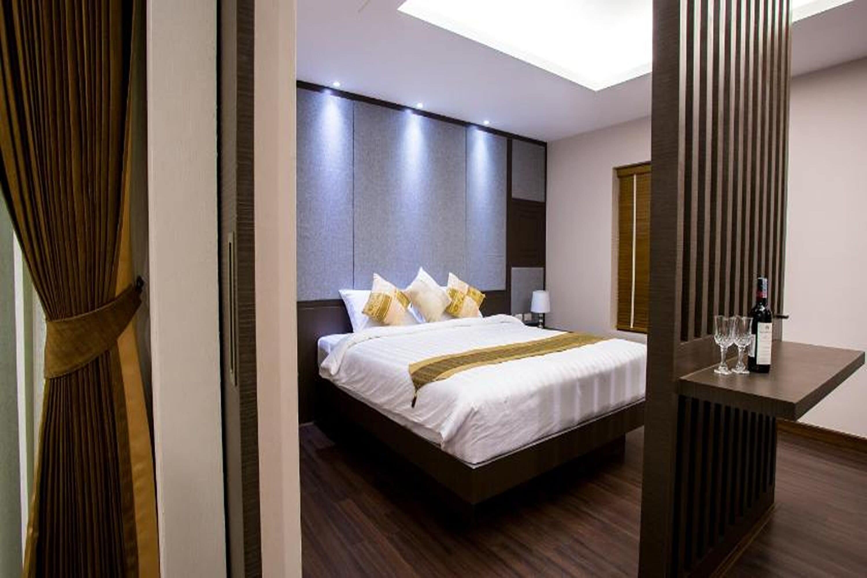 A modern hotel room with a large bed, a bedside table with a bottle and glasses, and a view of the sky through a skylight.