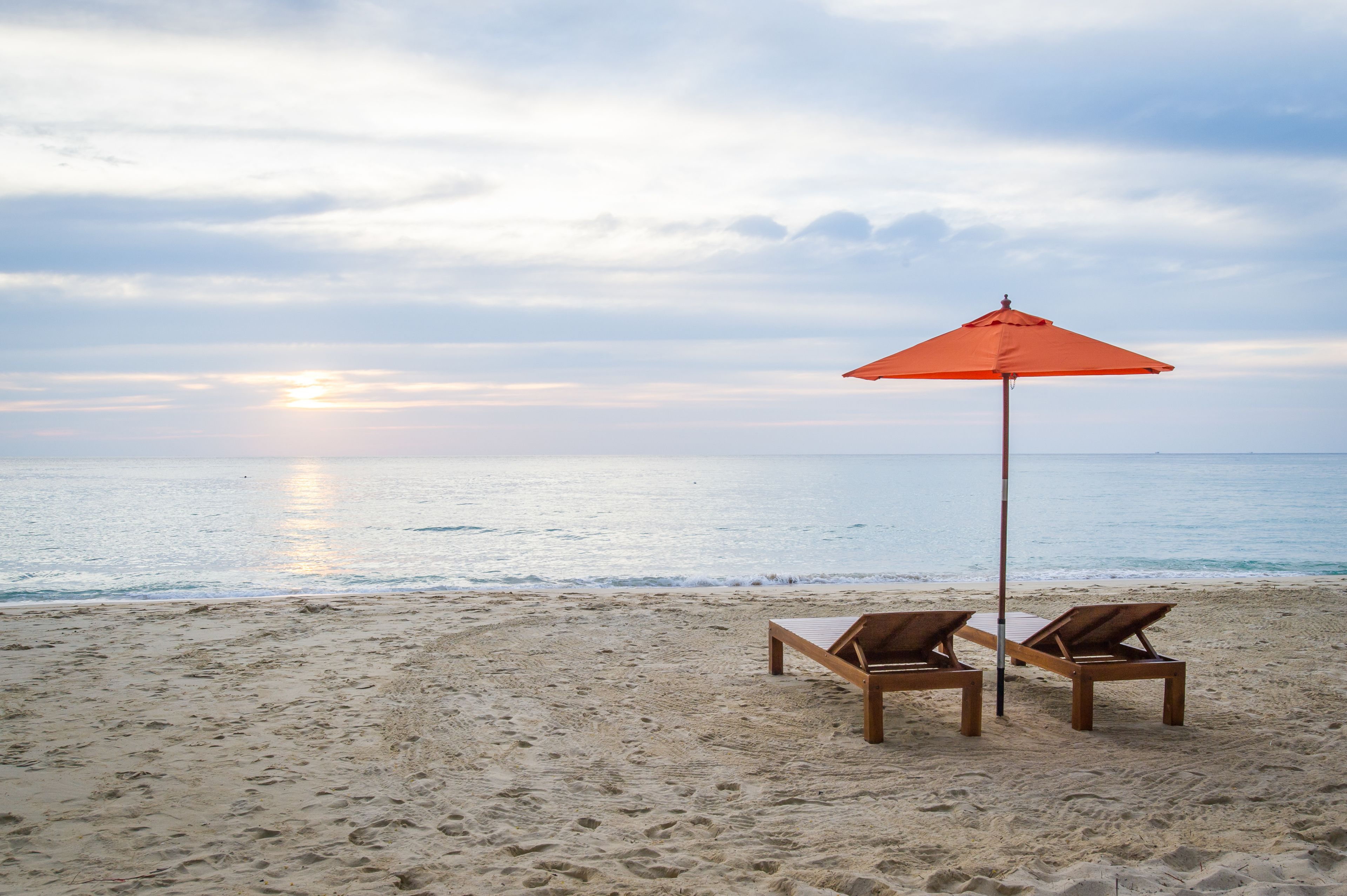 A beach with two wooden lounge chairs and an orange umbrella.