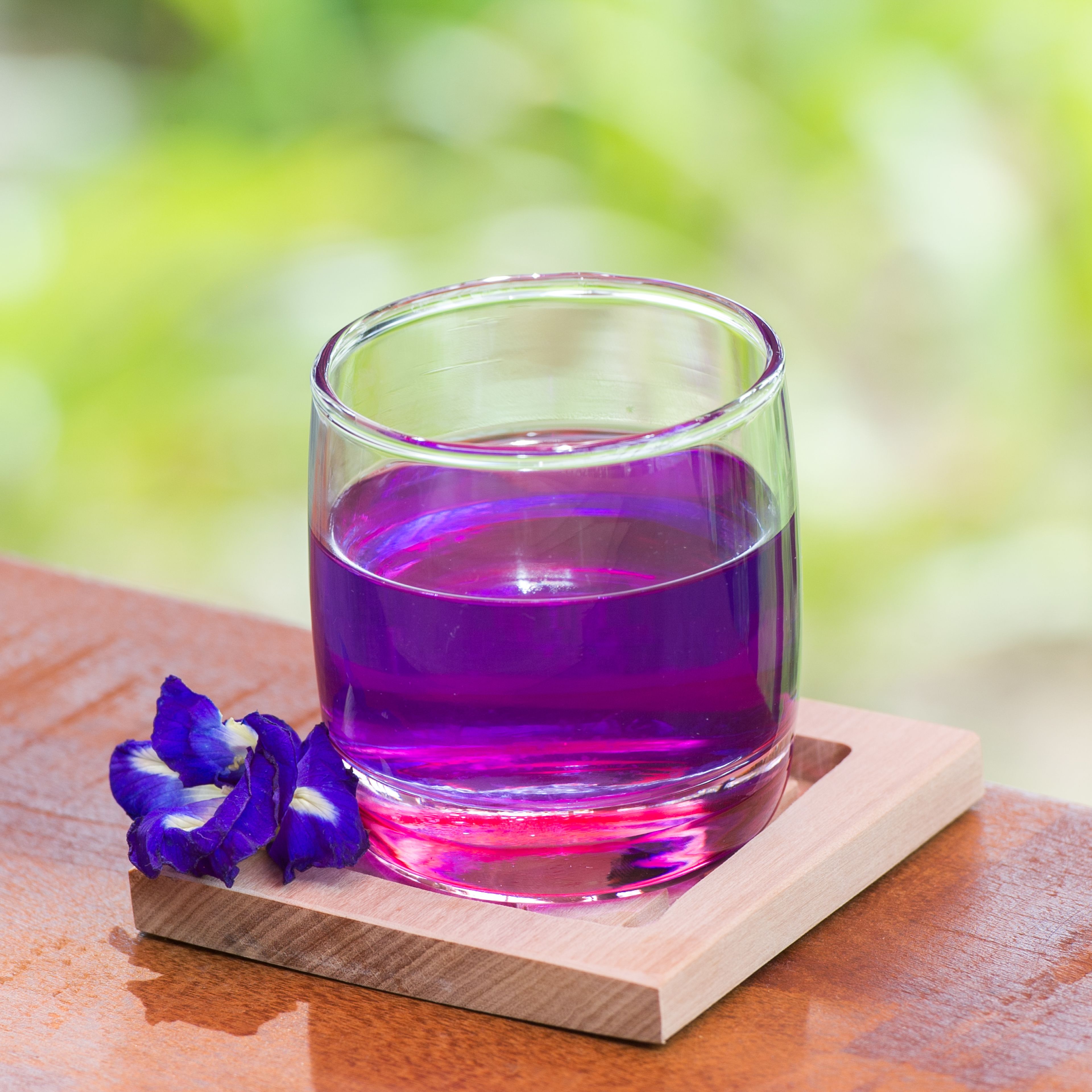 A glass of purple liquid on a wooden coaster with a flower beside it.