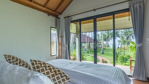 A bedroom with a large bed, a view of a tropical garden, and a sliding glass door leading to an outdoor area.
