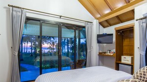 A hotel room with a large sliding glass door leading to a balcony with a view of palm trees and a sunset. There is a bed with a patterned blanket, a small TV on the wall, and a wooden cabinet with a microwave. - Medee Resort (Ko Kood)