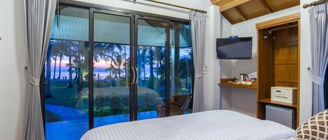 A hotel room with a large sliding glass door leading to a balcony with a view of palm trees and a sunset. There is a bed with a patterned blanket, a small TV on the wall, and a wooden cabinet with a microwave.