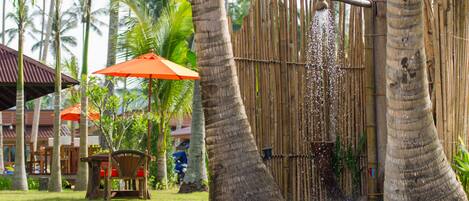 A bamboo shower enclosure with a water spout, surrounded by palm trees and a wooden fence.
