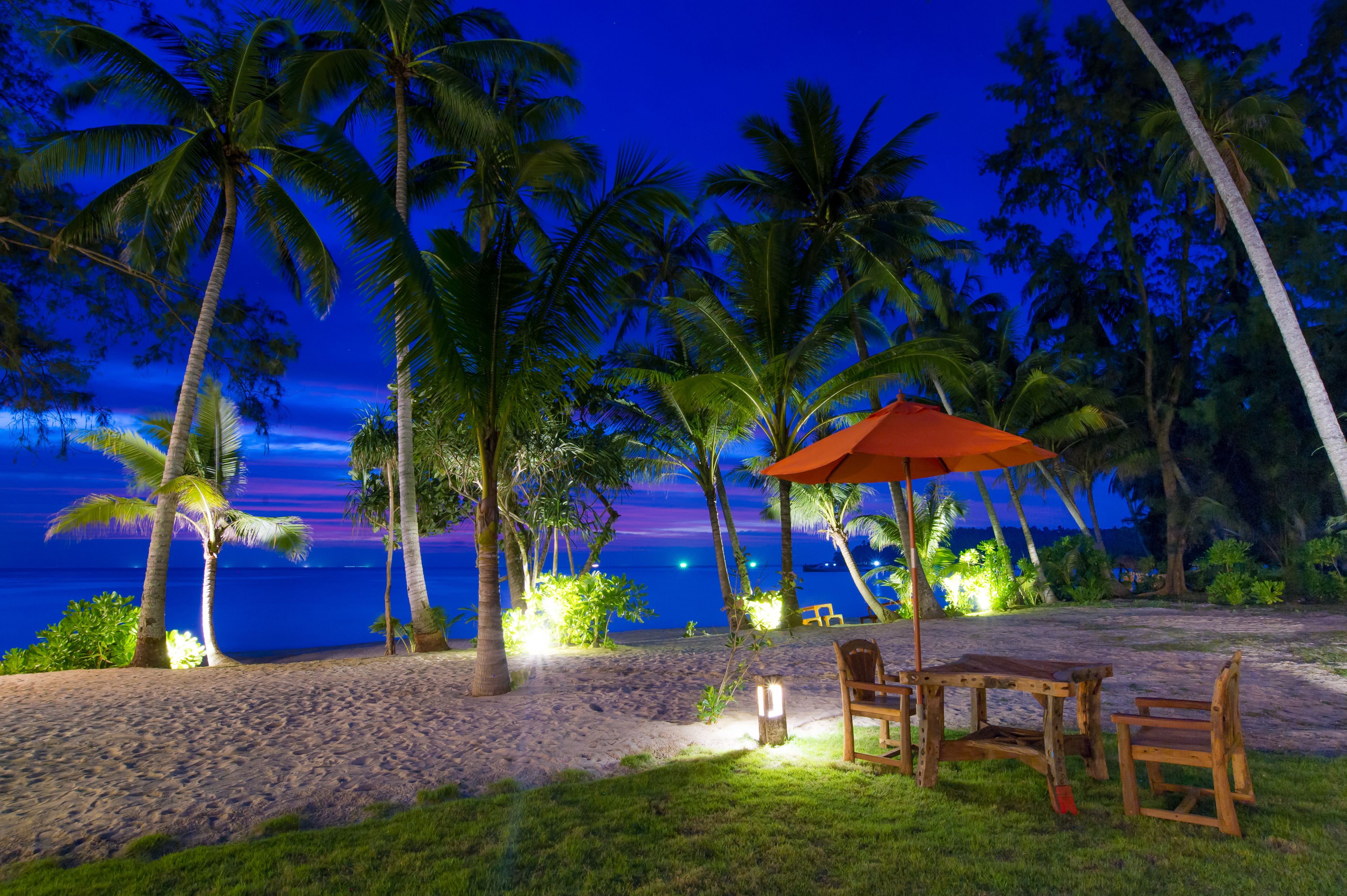 Escena de playa con palmeras, una mesa de madera con sillas y una sombrilla.