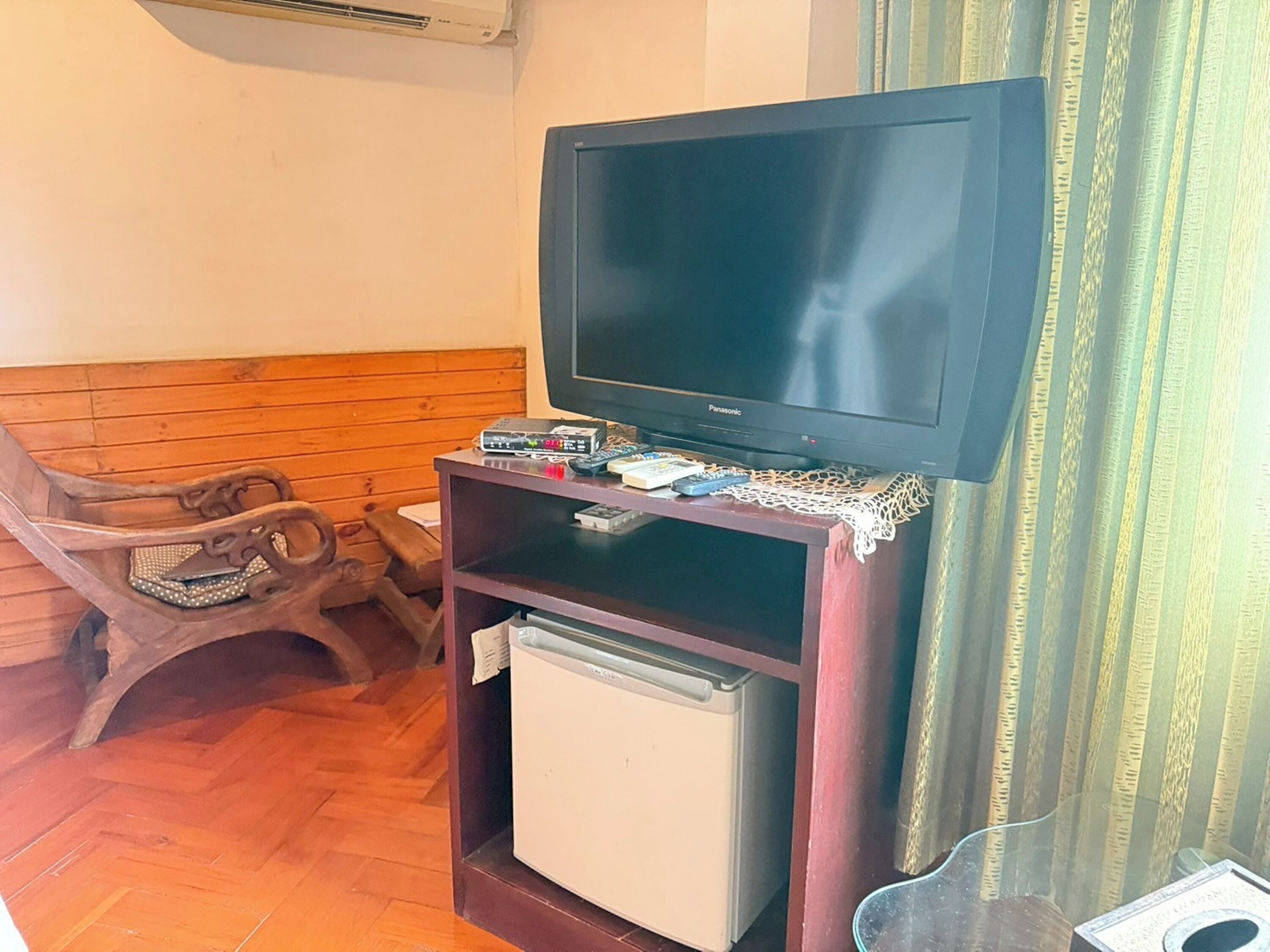 A small living area with a television on a wooden stand, a small refrigerator, and a wooden chair.