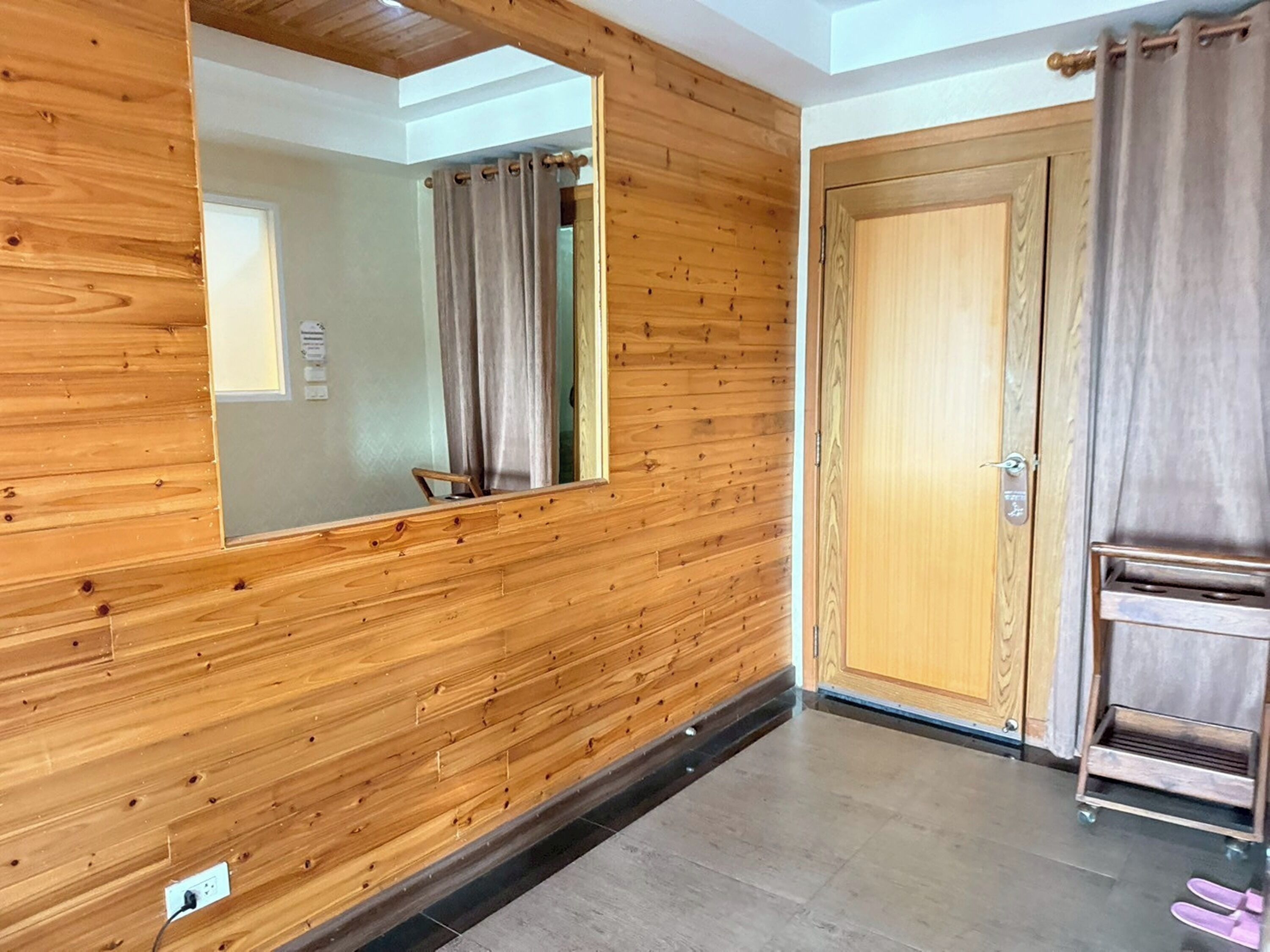 A room with wooden paneling, a large mirror, a door with a handle, and a small cart with slippers.