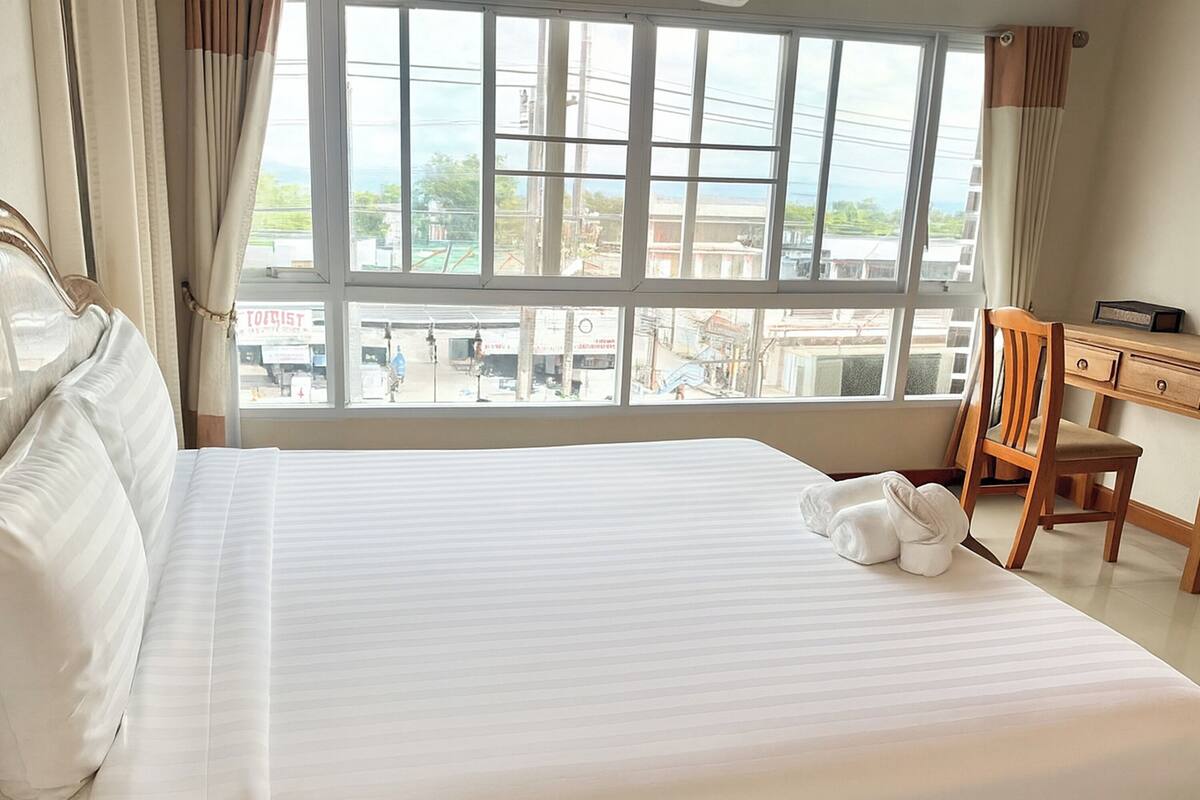 Habitación de hotel con una cama grande, una silla de madera, un escritorio y vistas de la ciudad a través de amplias ventanas.