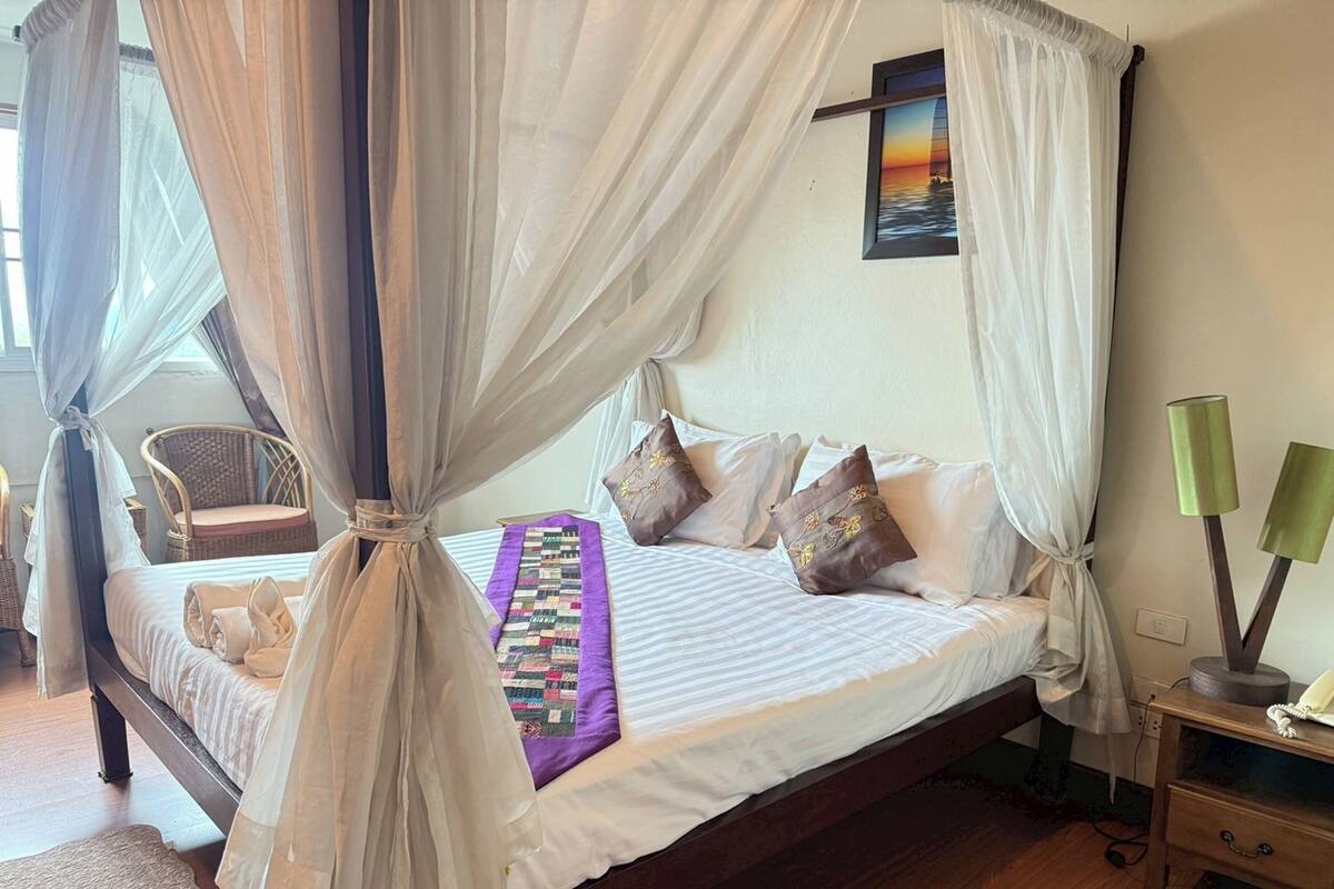 Una cama con dosel, ropa de cama blanca, un colcha morada y almohadas decorativas.