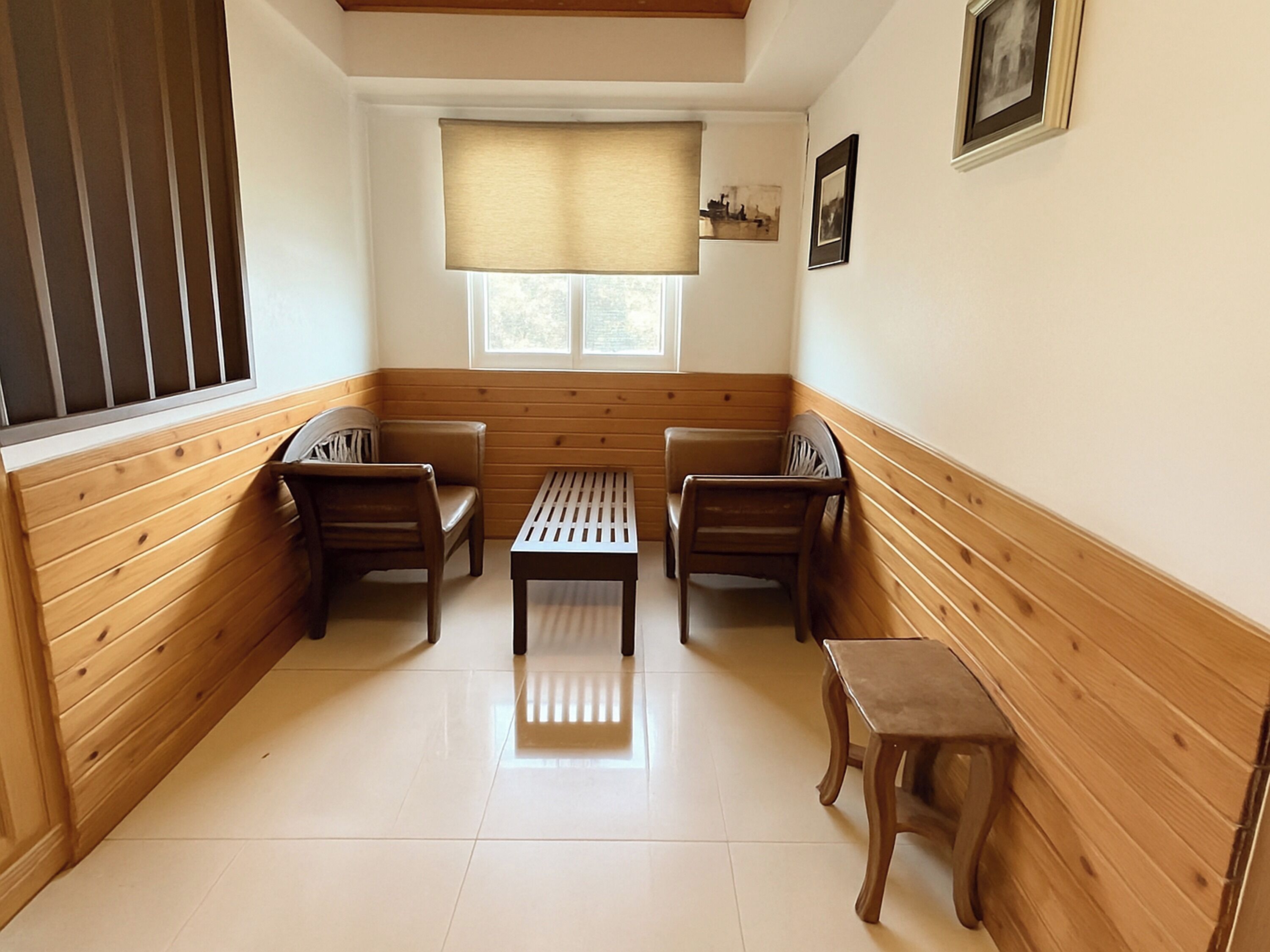 A corner room with wooden paneling, a small table, two chairs, a bench, and a window with a blind.