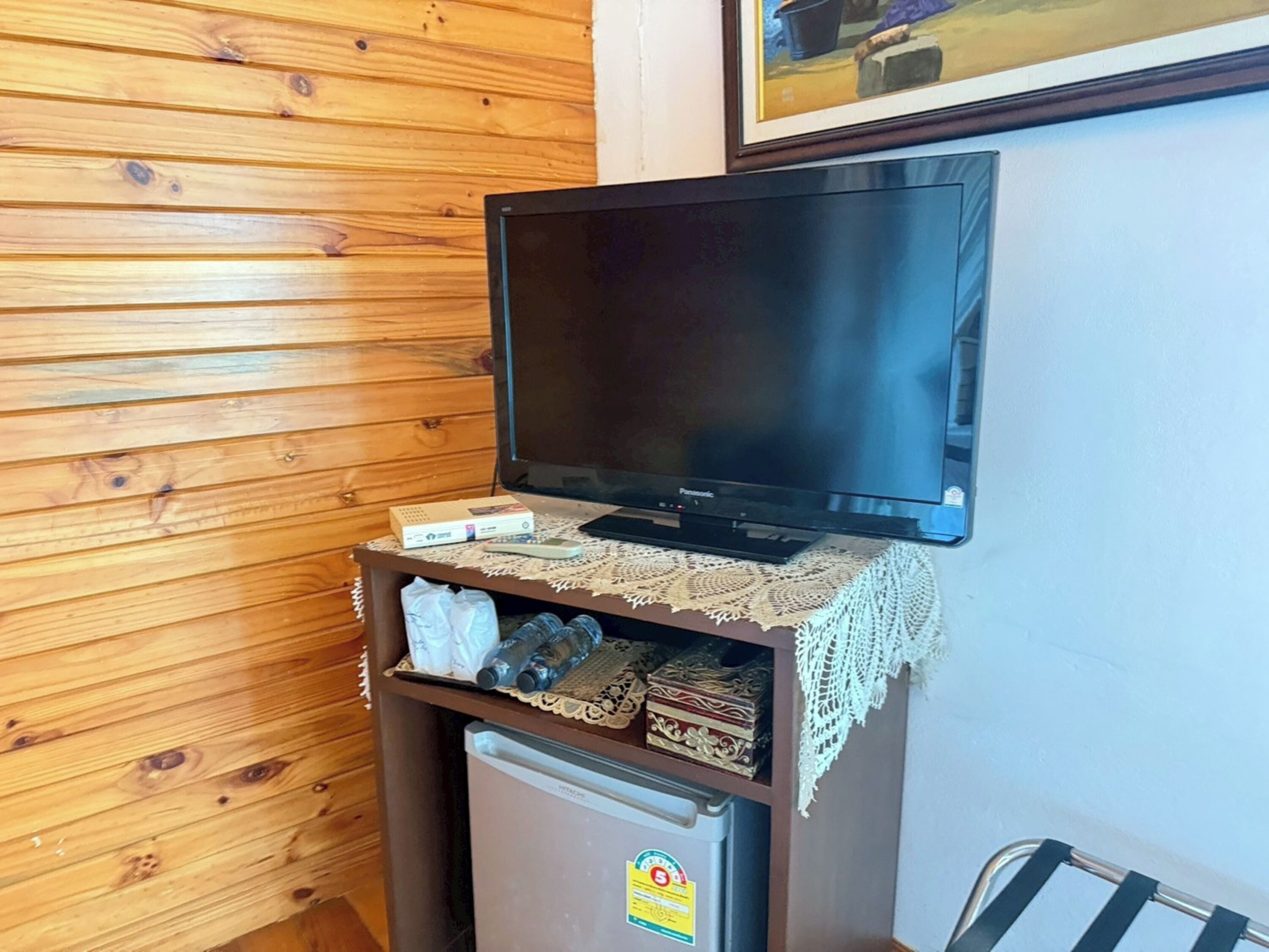 A wooden wall with a television on a wooden cabinet, a refrigerator below, and a framed picture above.