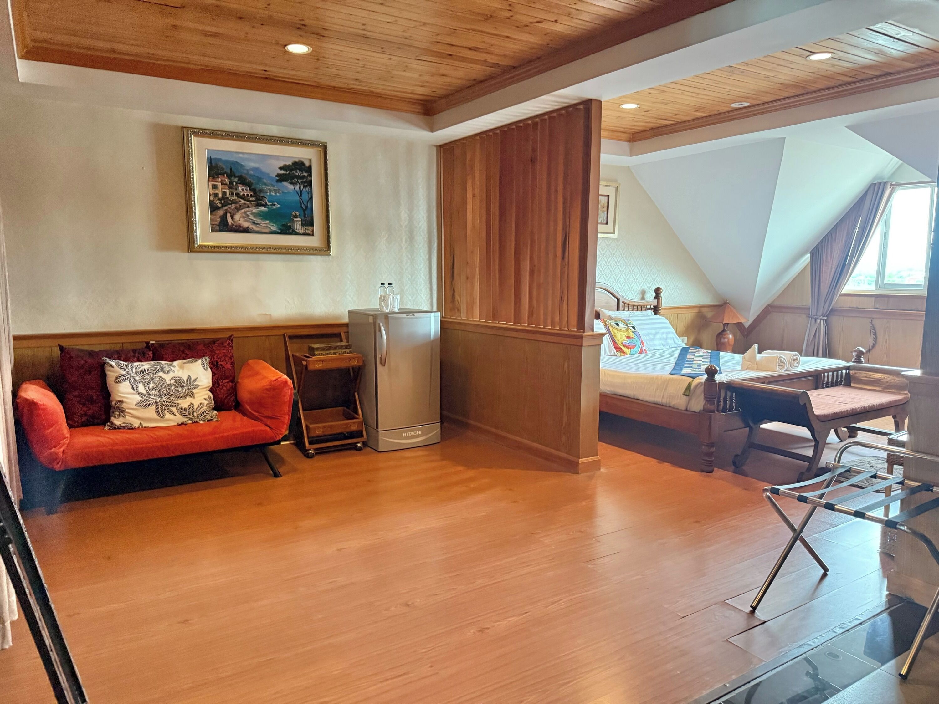 Ein gemütliches Zimmer mit Holzbalkendecke, einem roten Sofa, einer kleinen Kommode, einem Bett mit Blick und einem Glastisch.
