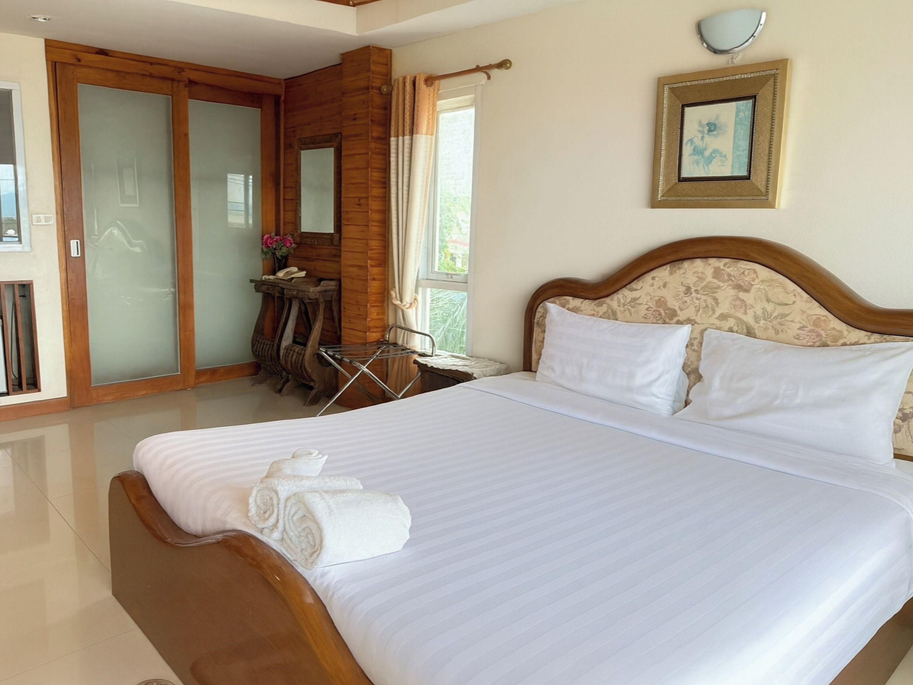 A hotel room with a large bed, wooden headboard, a desk with a chair, a small table with a vase of flowers, and a sliding glass door leading to a balcony.