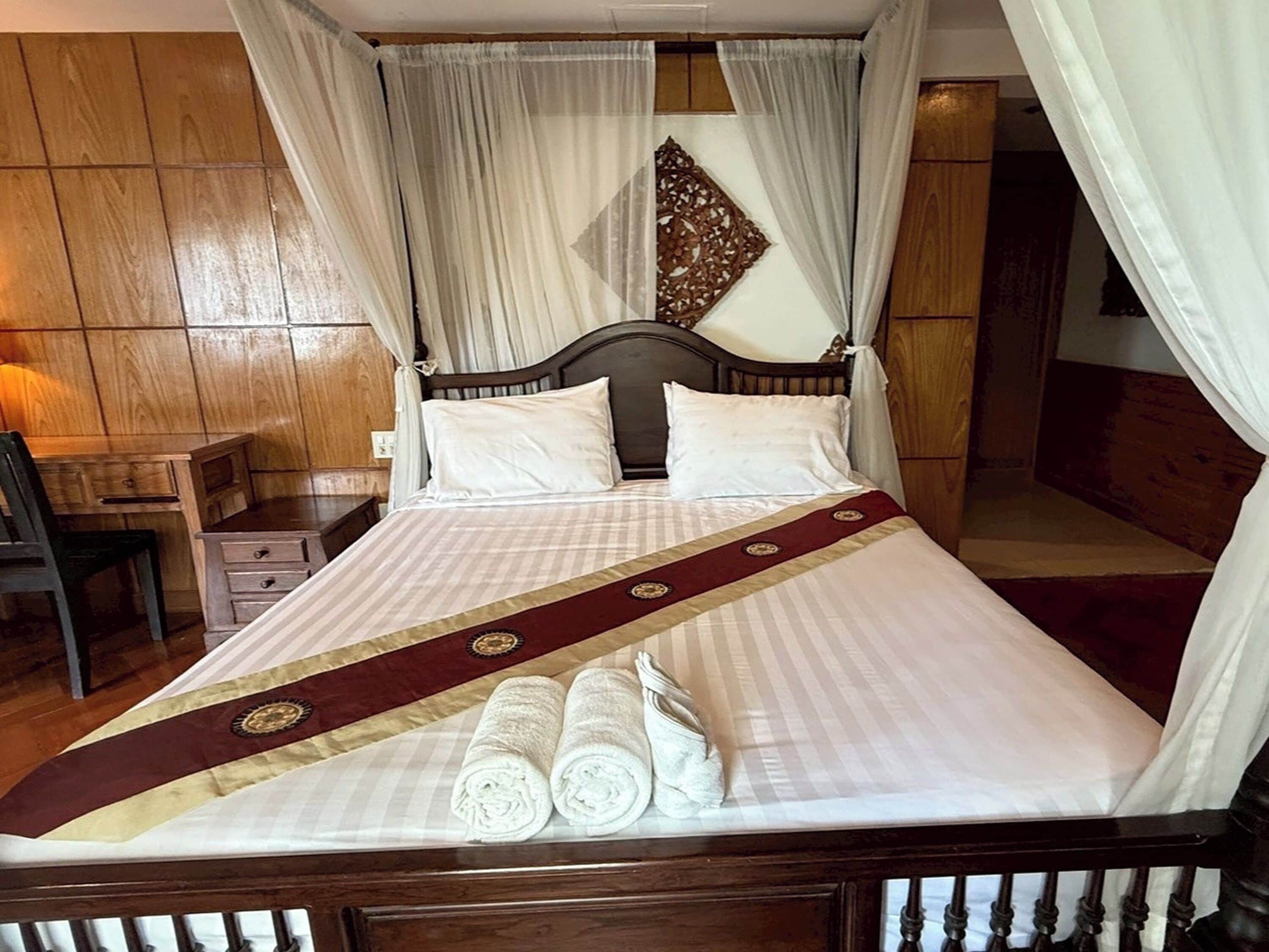 A neatly made bed with white linens and a red decorative runner, a wooden nightstand with a lamp, and a chair.