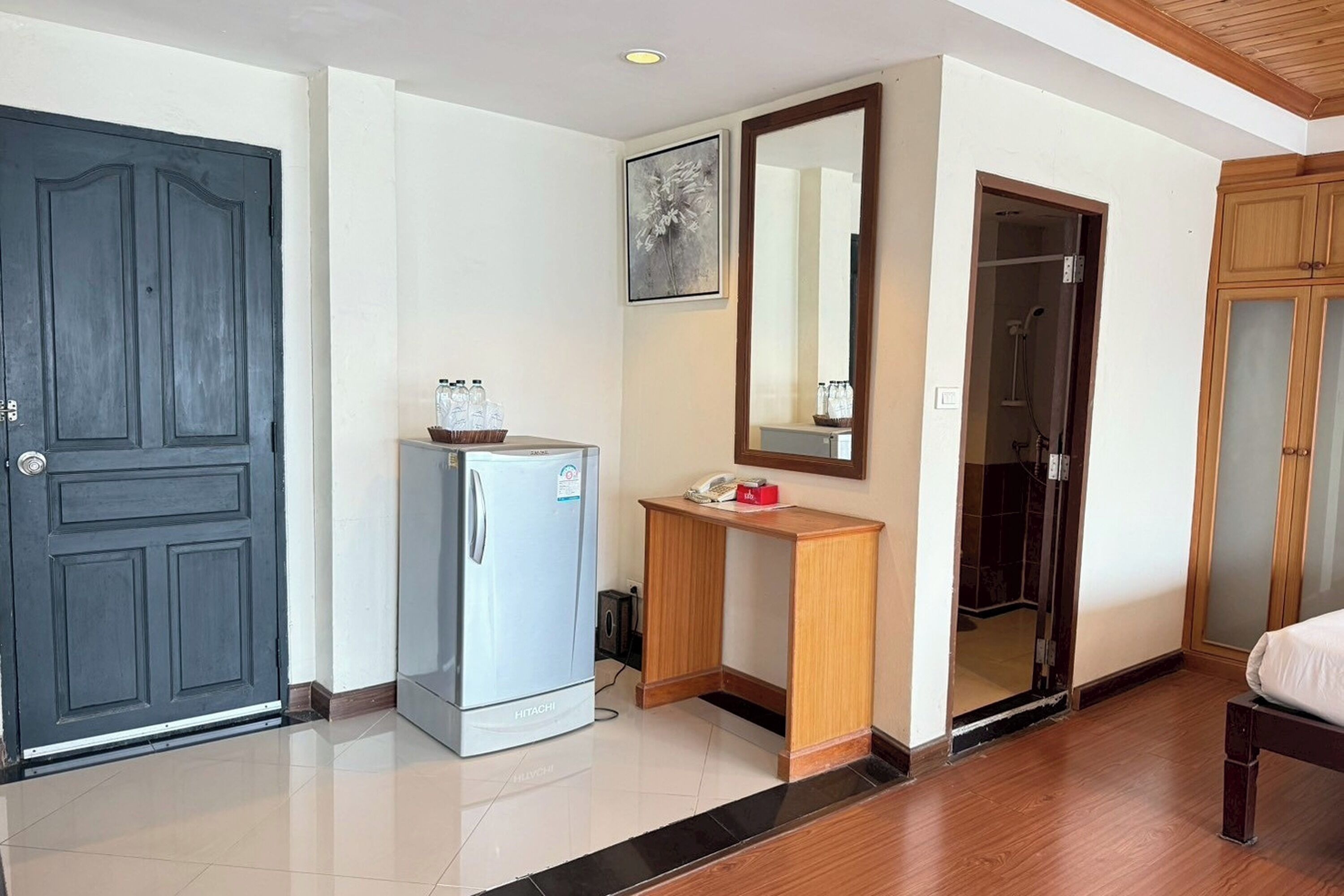 A hotel room with a dark wooden door, a Hitachi refrigerator, a wooden console table with a telephone, a mirror, and a shower area.