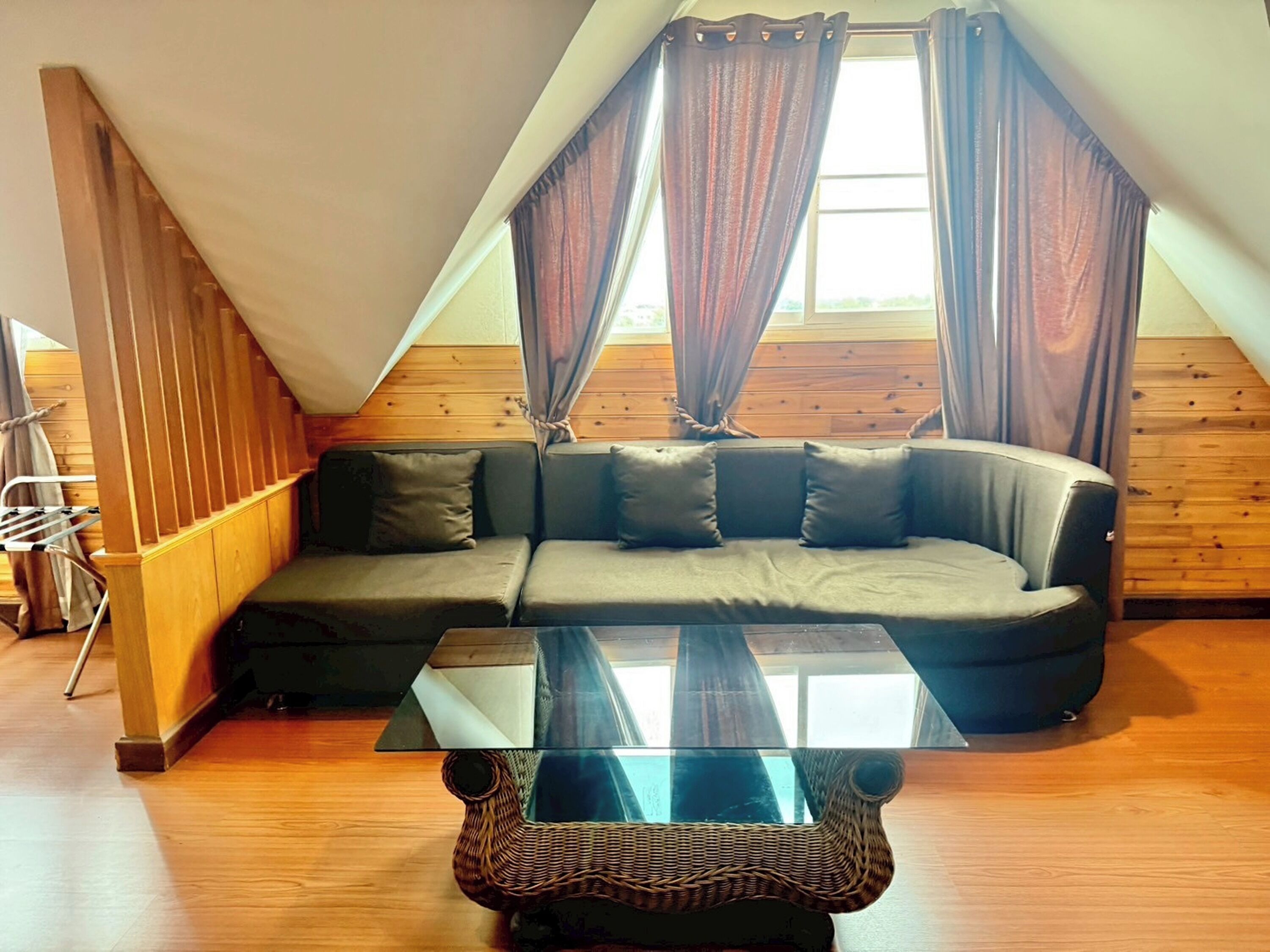 Ein gemütliches Zimmer mit Holzbalkendecke, einem Ecksofa, einem Glastisch und einem Fenster mit Vorhängen.