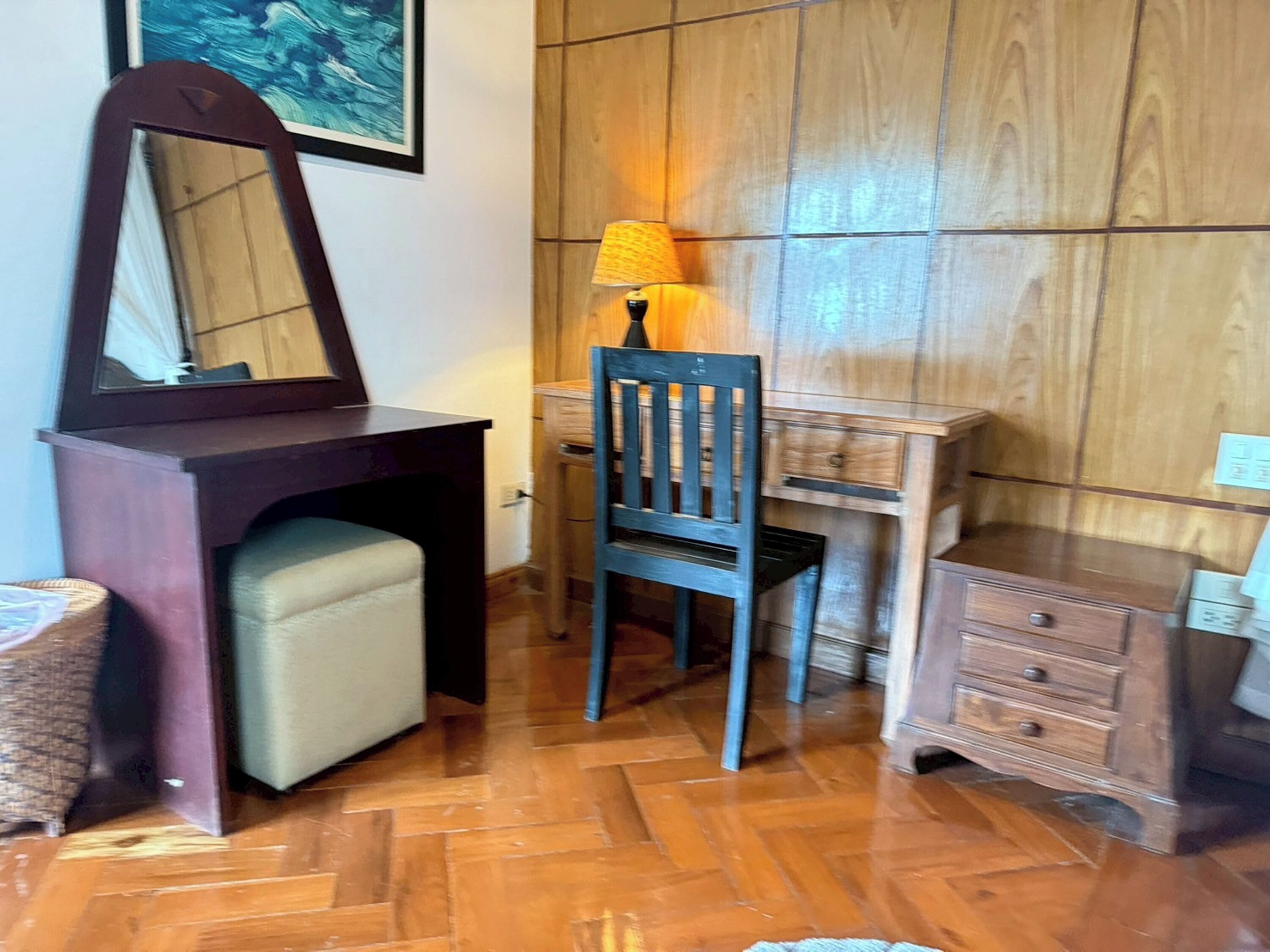 Una habitació amb escriptori de fusta, cadira, mirall, làmpada i un petit armari.