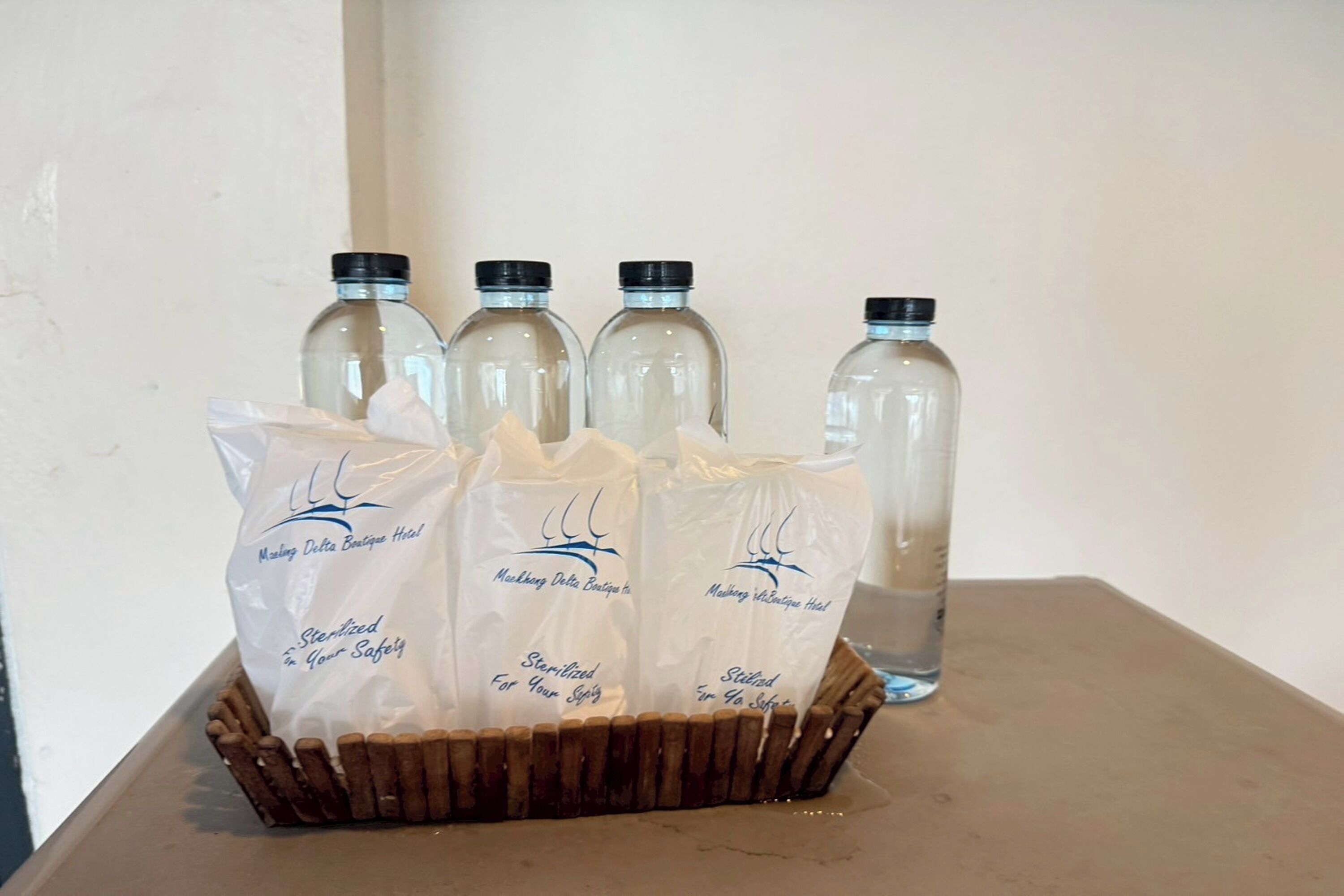 A wicker basket containing five white bags with text, and four clear glass bottles with black caps in the background.