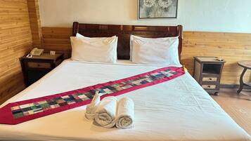 A neatly made bed with a quilt, pillows, and rolled towels. There is a wooden headboard, a bedside table with a phone, and a small table with a lamp.