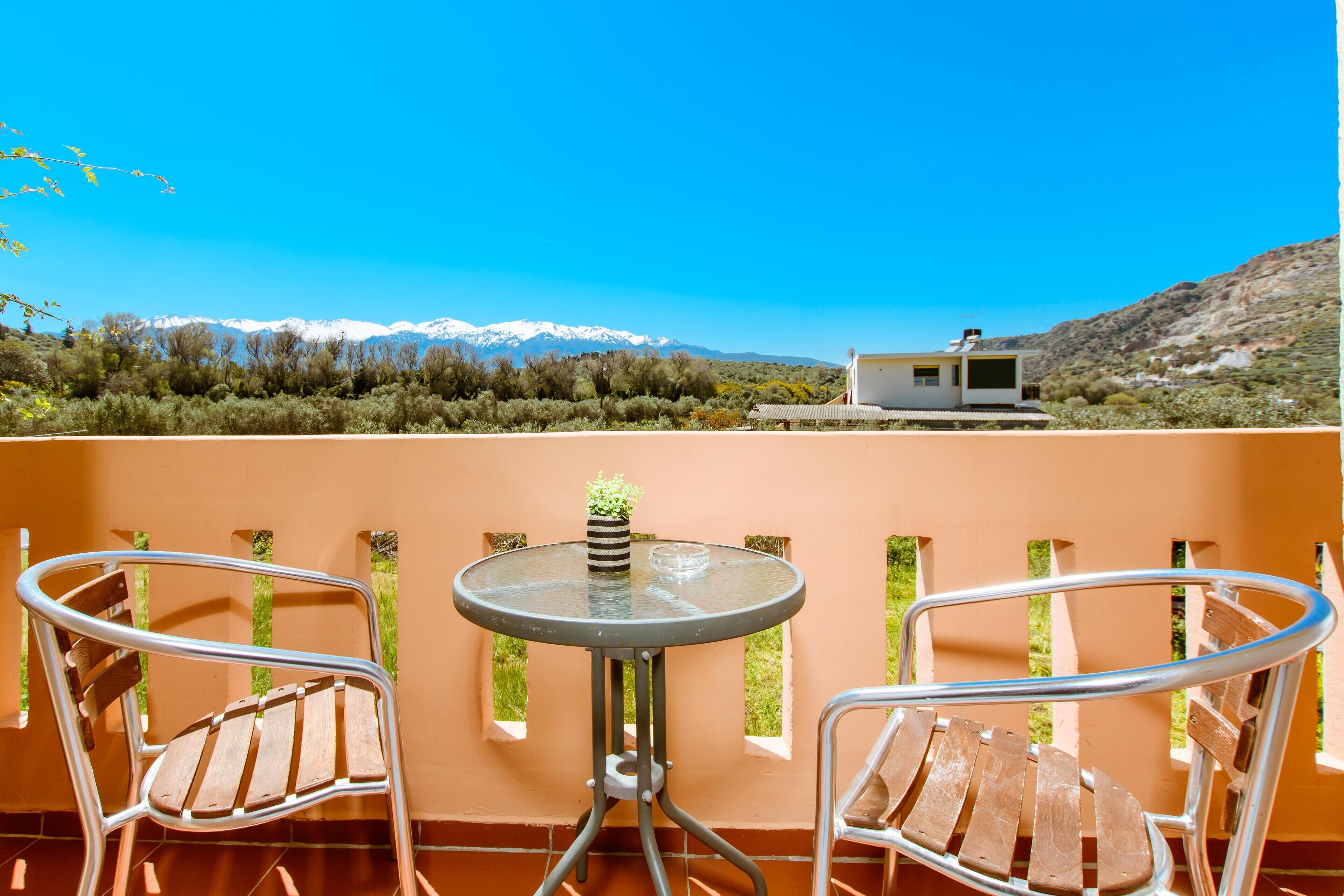A balcony with two metal chairs and a small table, overlooking a mountainous landscape with a cabin.