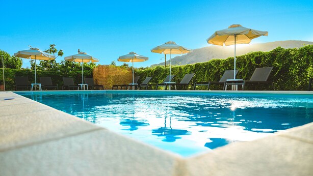 A swimming pool with lounge chairs and umbrellas, surrounded by greenery and mountains. - Koulouris Apartments (Apokoronas)