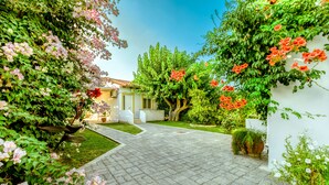 A paved courtyard with a pathway leading to a building, surrounded by lush greenery and flowering plants. - Koulouris Apartments (Apokoronas)