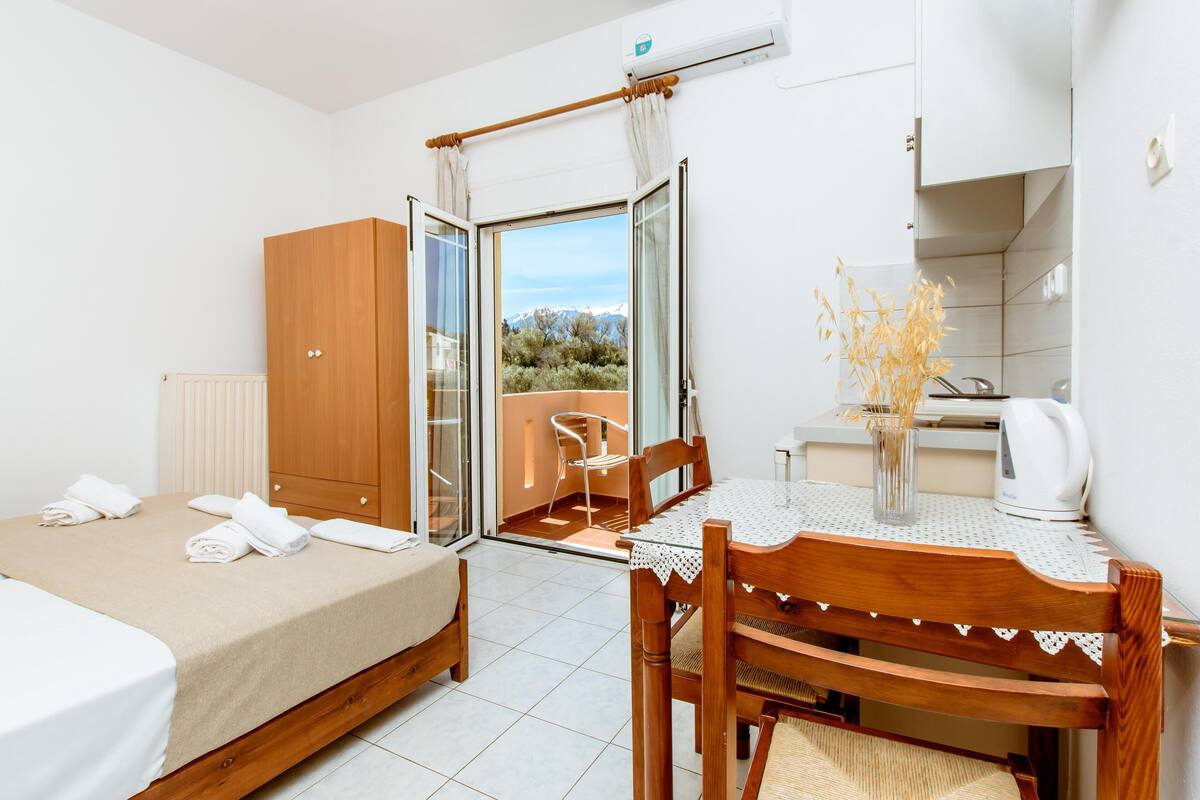 A hotel room with a bed, a table with chairs, a view of the outdoors, and a kitchenette.