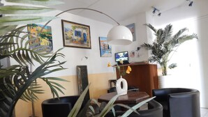 Um quarto com um balcão de recepção de madeira, uma grande planta em vaso, um vaso branco com flores e um quadro na parede.