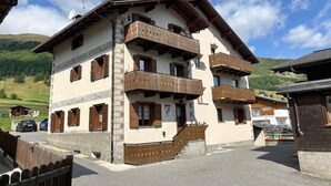 A multi-story building with wooden balconies, a stone facade, and a mountainous backdrop. - Appartamento Baita Cusini Saroch (Livigno)