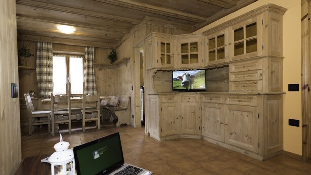 A wooden interior with a laptop on a table, a TV mounted on a cabinet, and a dining area with chairs and a table. - Appartamento Baita Cusini Saroch (Livigno)