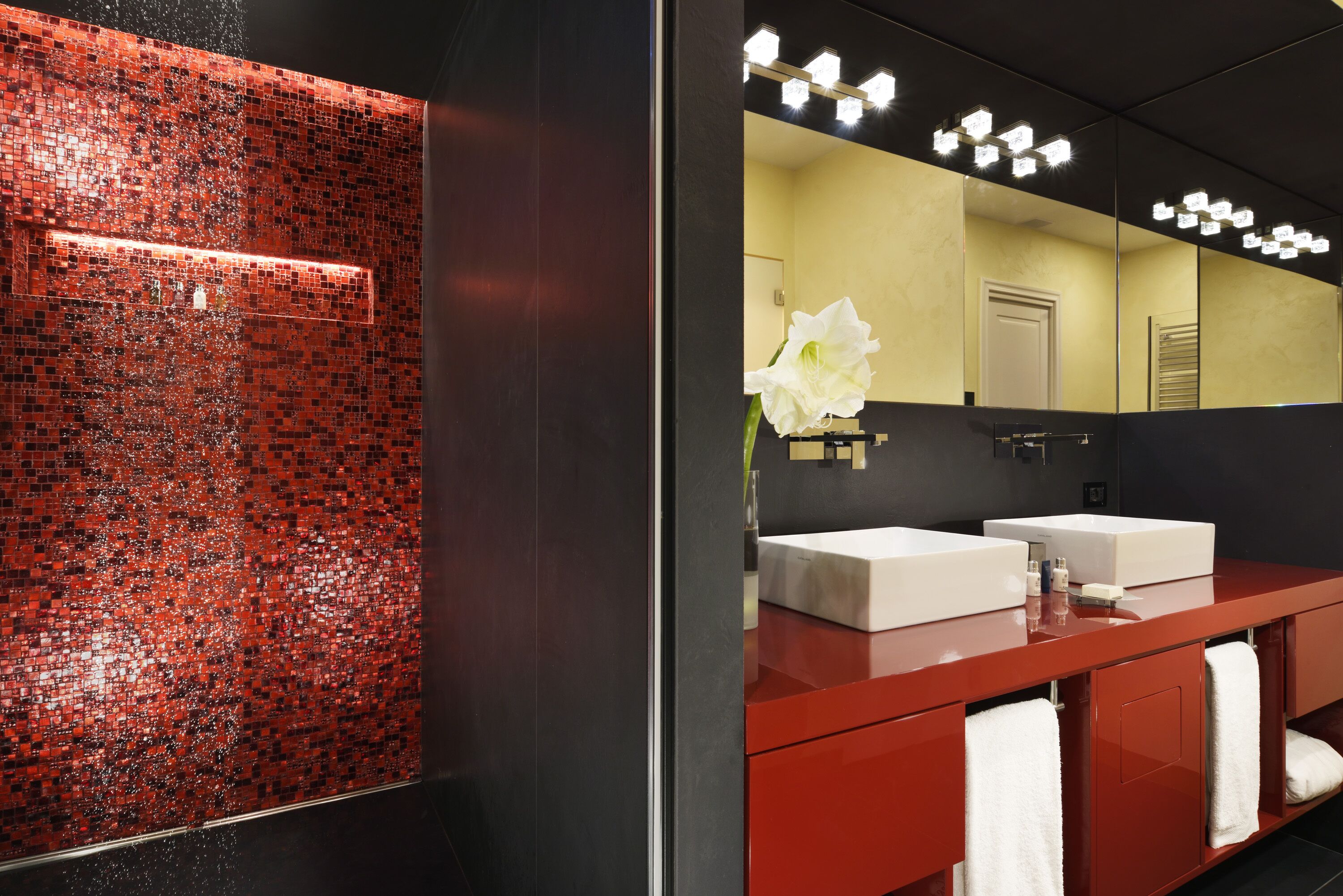 A modern bathroom with a red and black tiled wall, two sinks, and a large mirror with lights.