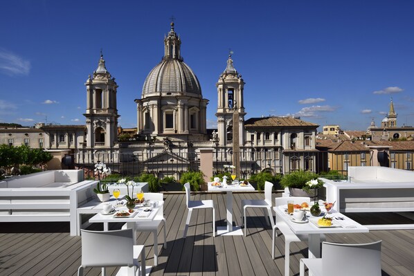 An outdoor dining area with white tables and chairs, a view of a historic building with domes and towers, and a clear blue sky. - Lifestyle Suites Rome (Rome)