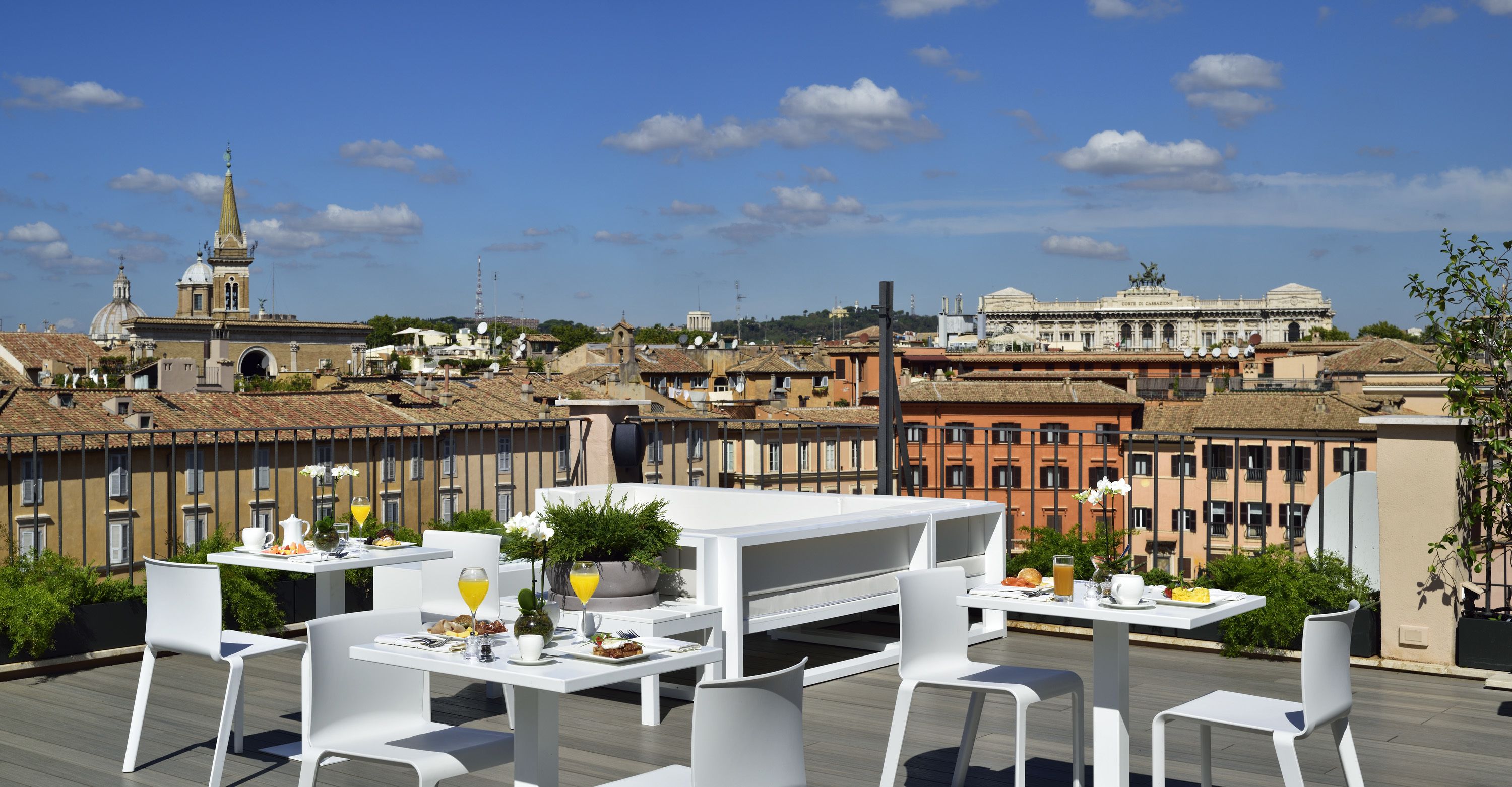 A rooftop terrace with white tables and chairs, a dining setup with plates, glasses, and a potted plant.
