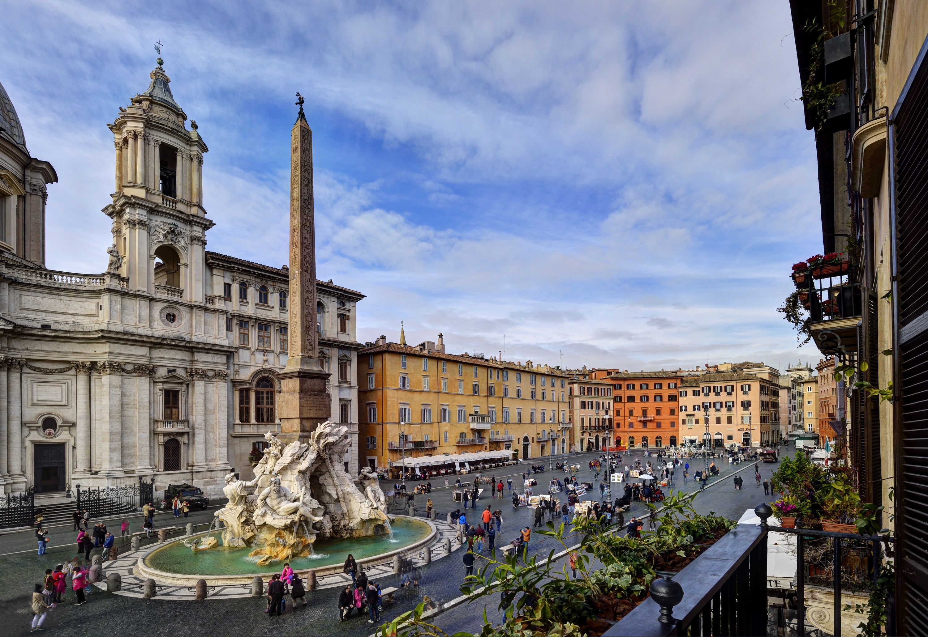 A public square with a historic fountain, a tall obelisk, and classical architecture.