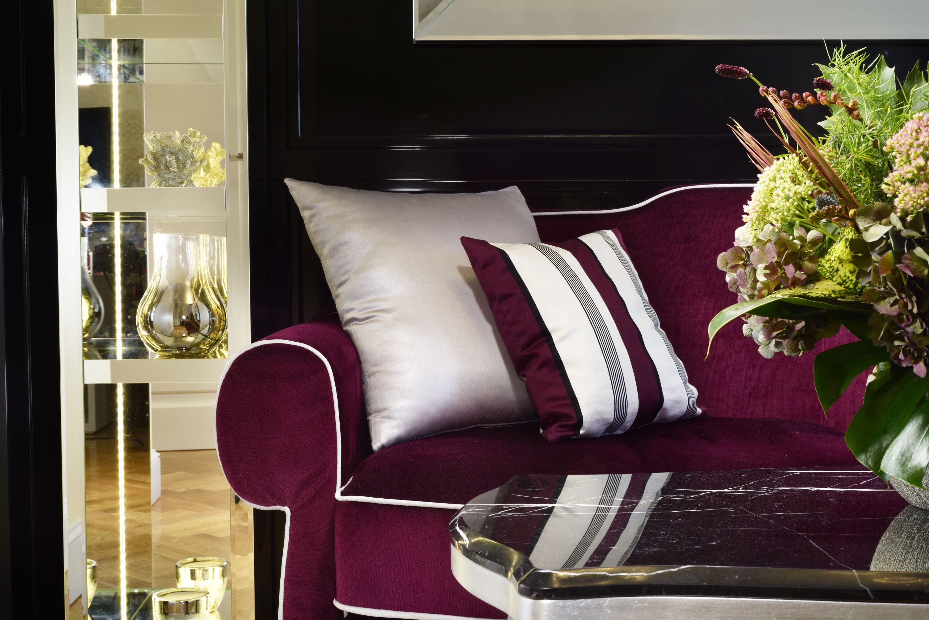 A dark-colored sofa with white and maroon cushions, a marble coffee table, and a floral arrangement.