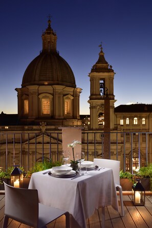 An outdoor dining area with a table set for two, a potted plant, and a lantern. In the background, there is a large dome and bell tower structure.