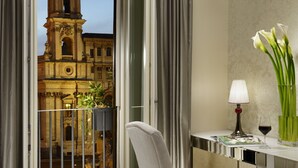 Uma varanda com vista para um edifício histórico, uma cadeira, uma mesa com abajur e um copo de vinho, e um vaso com flores.