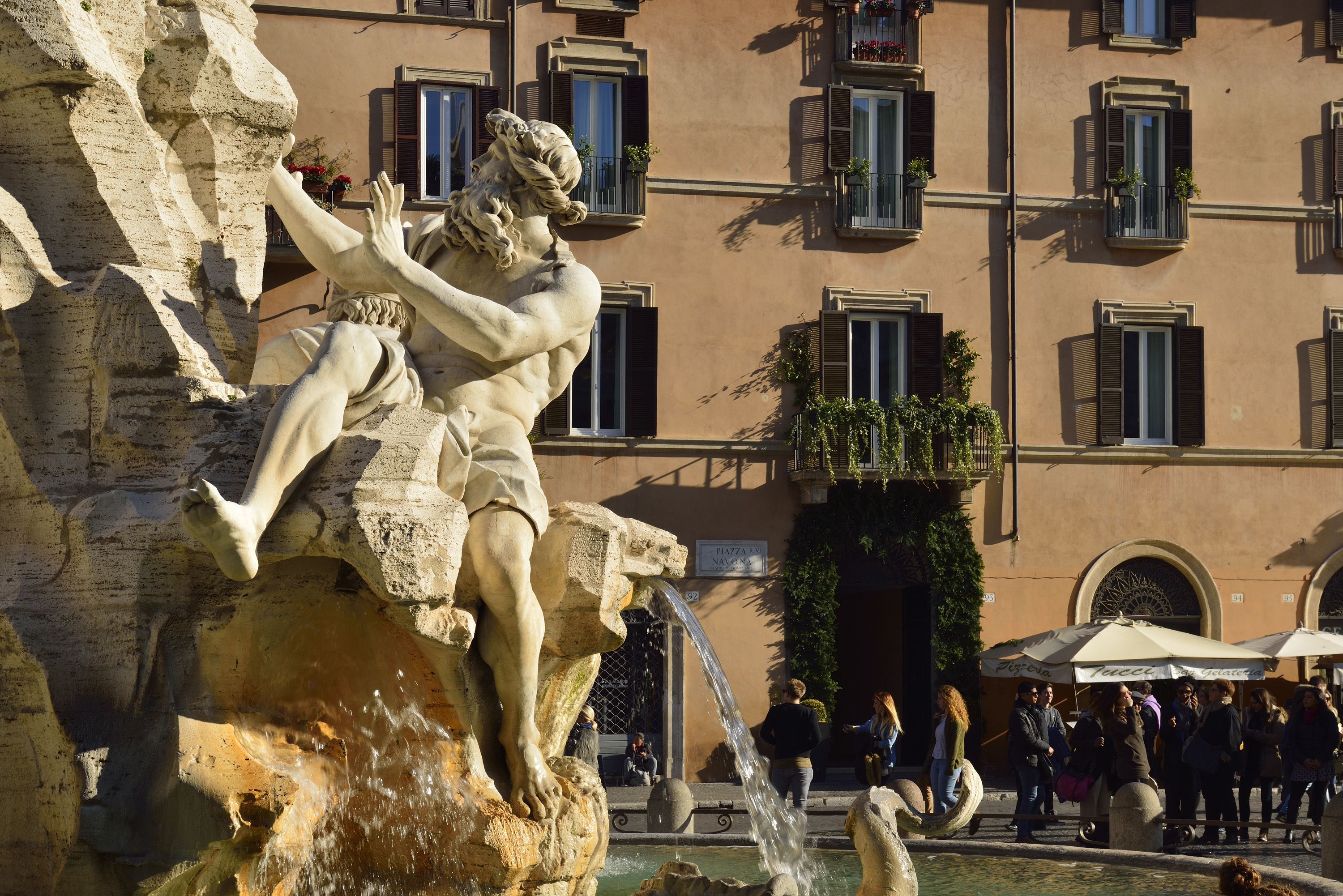 A stone fountain with a statue of a man reaching out, surrounded by a crowd of people.