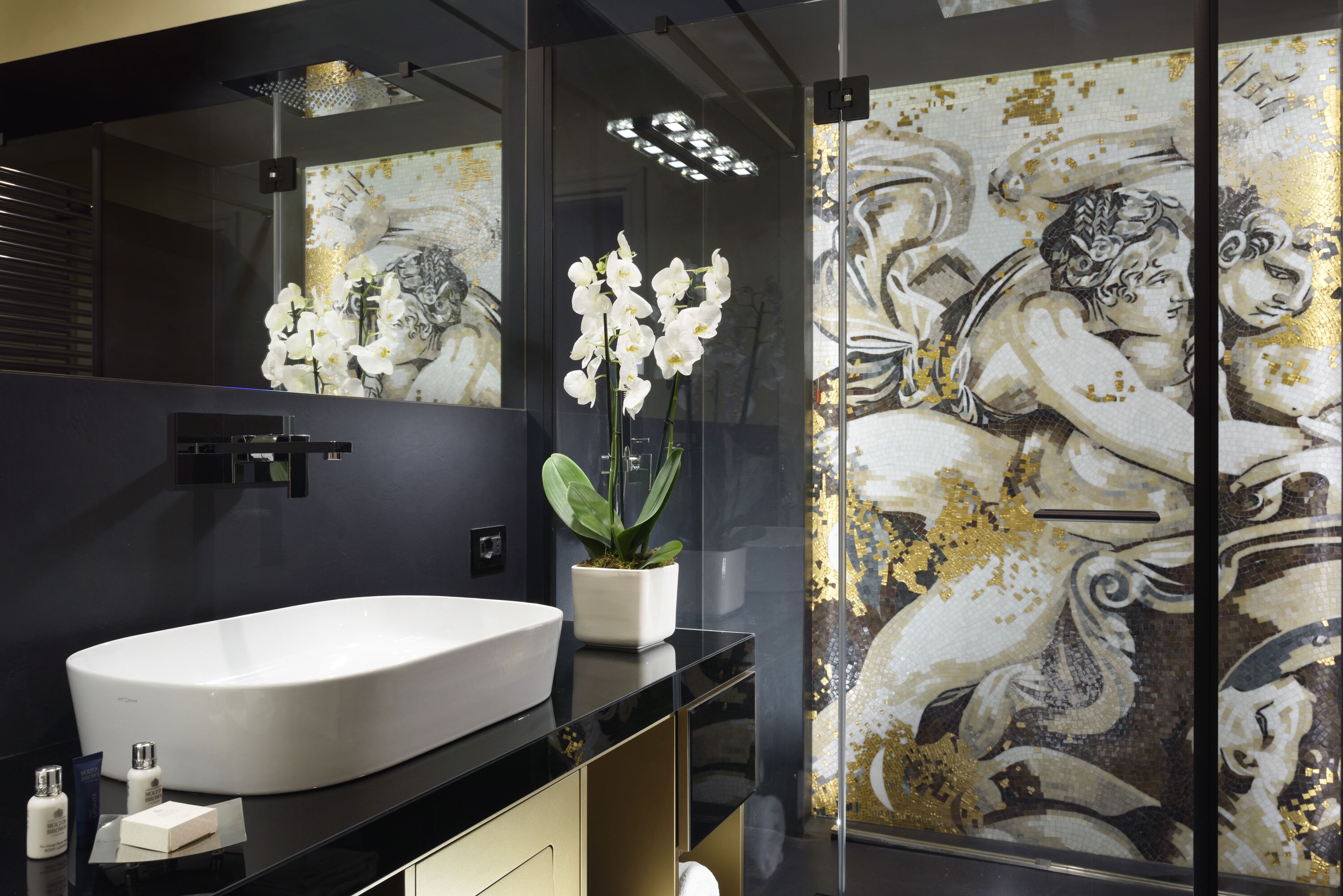 Casa de banho moderna com uma grande pia branca, uma orquídea em vaso e um painel decorativo com mosaico dourado e branco.