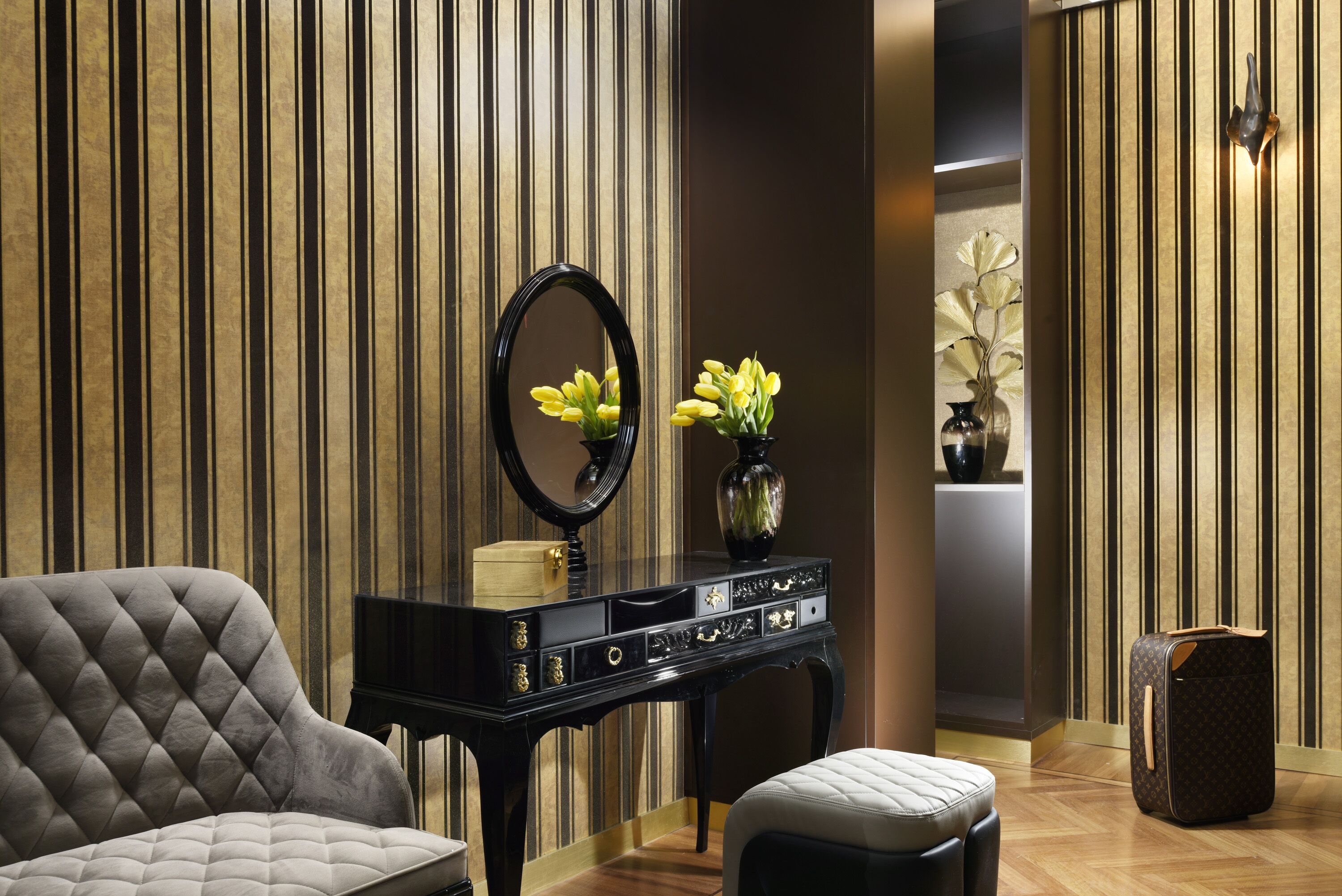 Una habitación con una consola negra, un sillón tapizado de gris, un banco blanco acolchado, un espejo negro y una maleta Louis Vuitton.