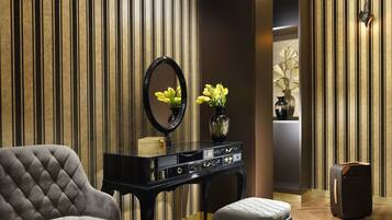 Una habitación con una consola negra, un sillón tapizado de gris, un banco blanco acolchado, un espejo negro y una maleta Louis Vuitton.