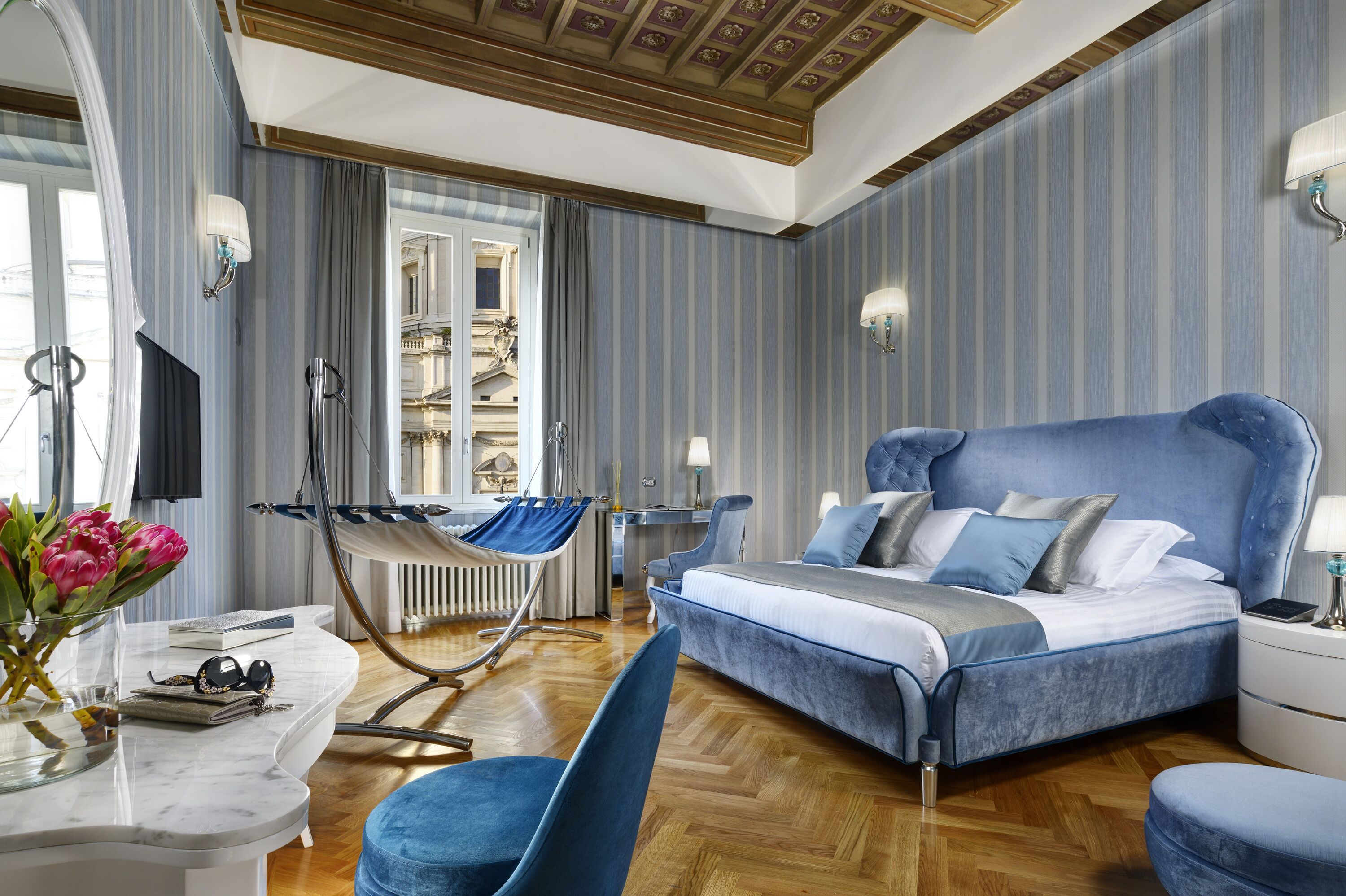 Een moderne hotelkamer met een groot bed, een zithoek met een stoel, een bureau en uitzicht op een gebouw door een raam.