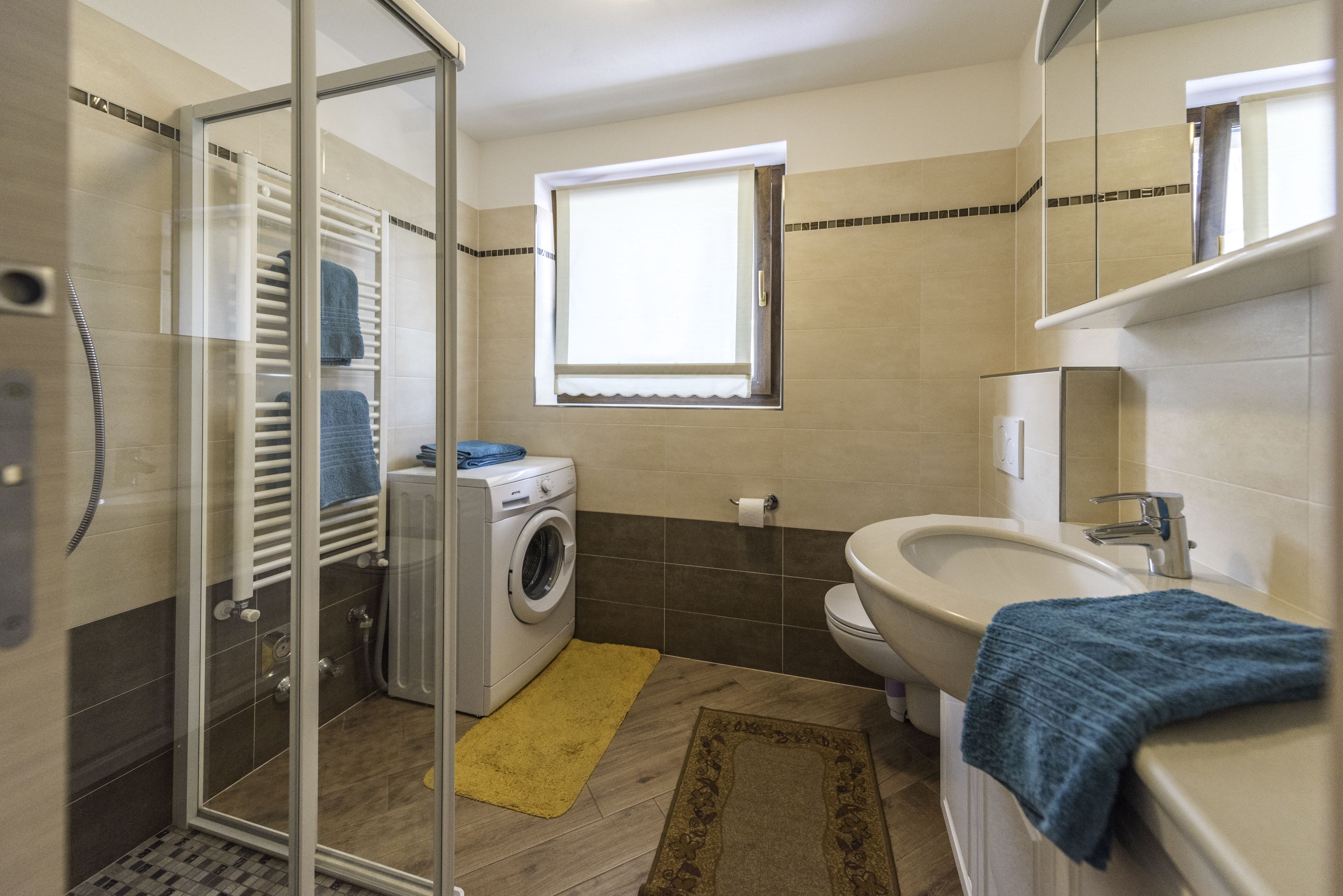 A bathroom with a glass shower enclosure, a washing machine, a sink, and a window.