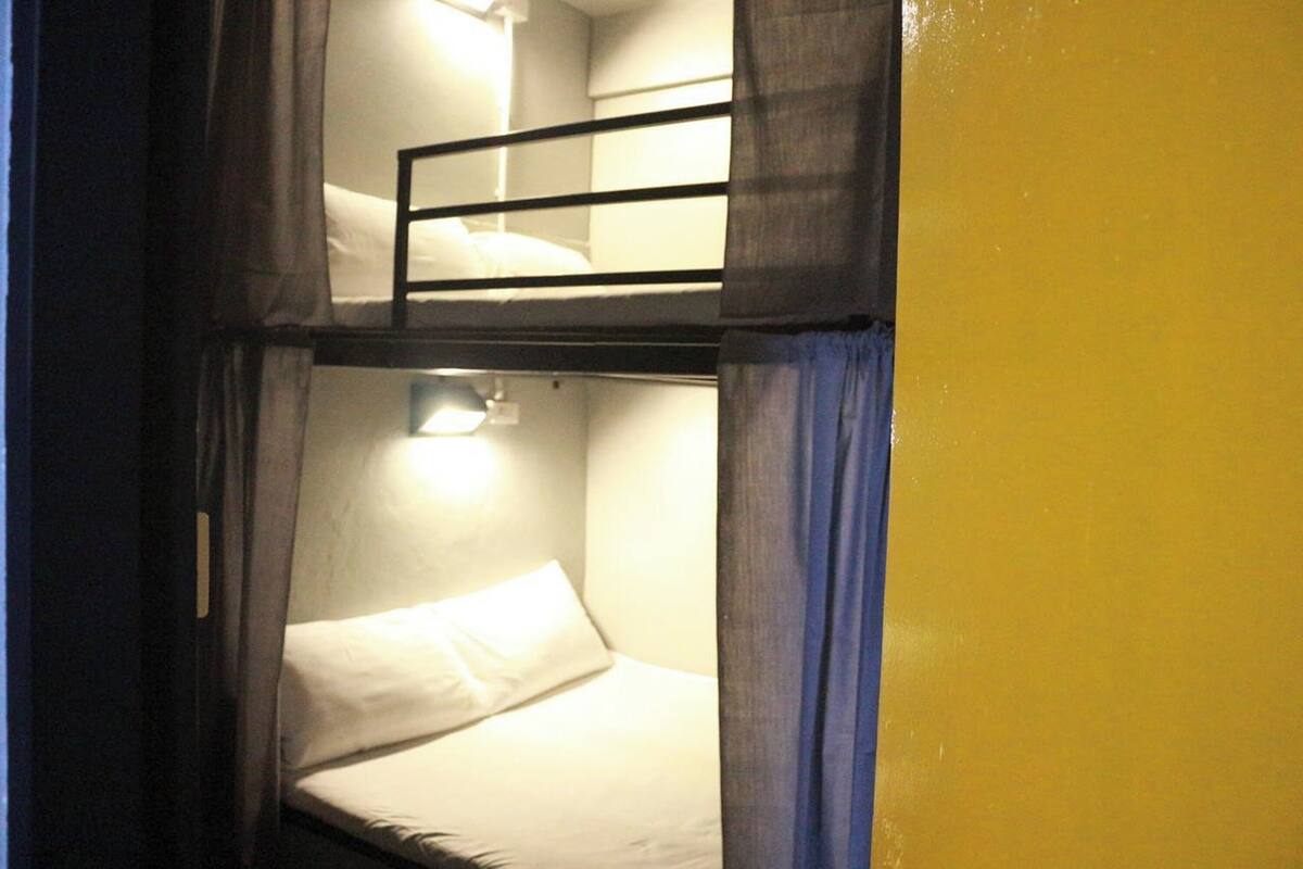 Habitación con literas, una cama individual, una mesita pequeña, cortinas y una lámpara fijada a la pared.