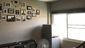 A small, well-lit room with a kitchenette, a dining area with a table and chairs, a refrigerator, and a wall adorned with framed pictures. - Hubb Hostel Phuket Airport (Sa Khu)